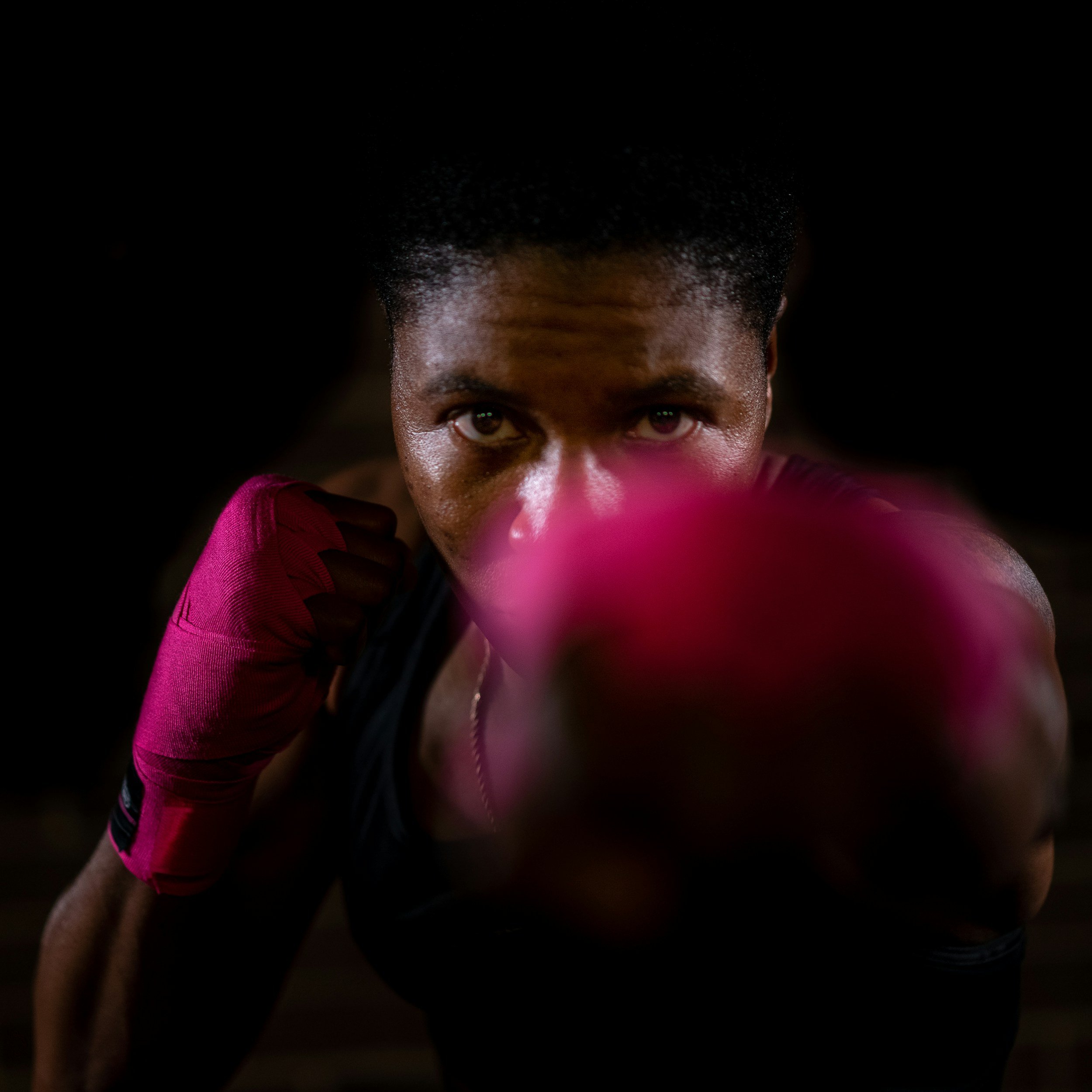 A person with dark skin and short natural hair holding a pink boxing glove close to their face, wearing a pink shirt.