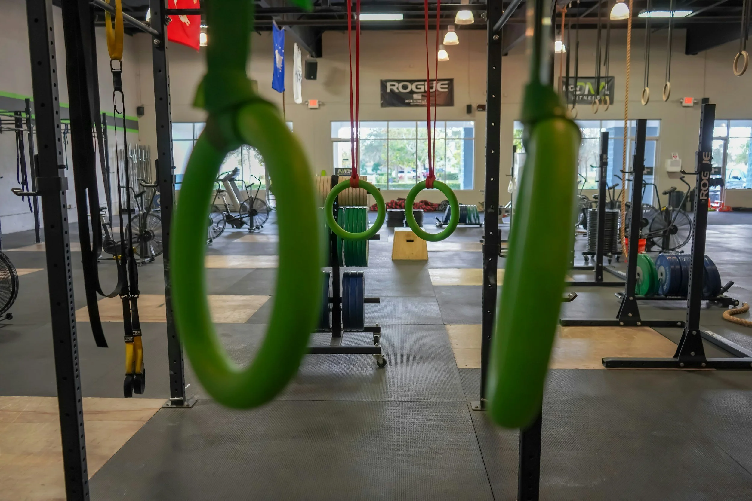 Interior of a gym with workout equipment, including green gymnastic rings hanging from red straps, and various exercise machines in the background. The space is well-lit with large windows and ceiling lights.