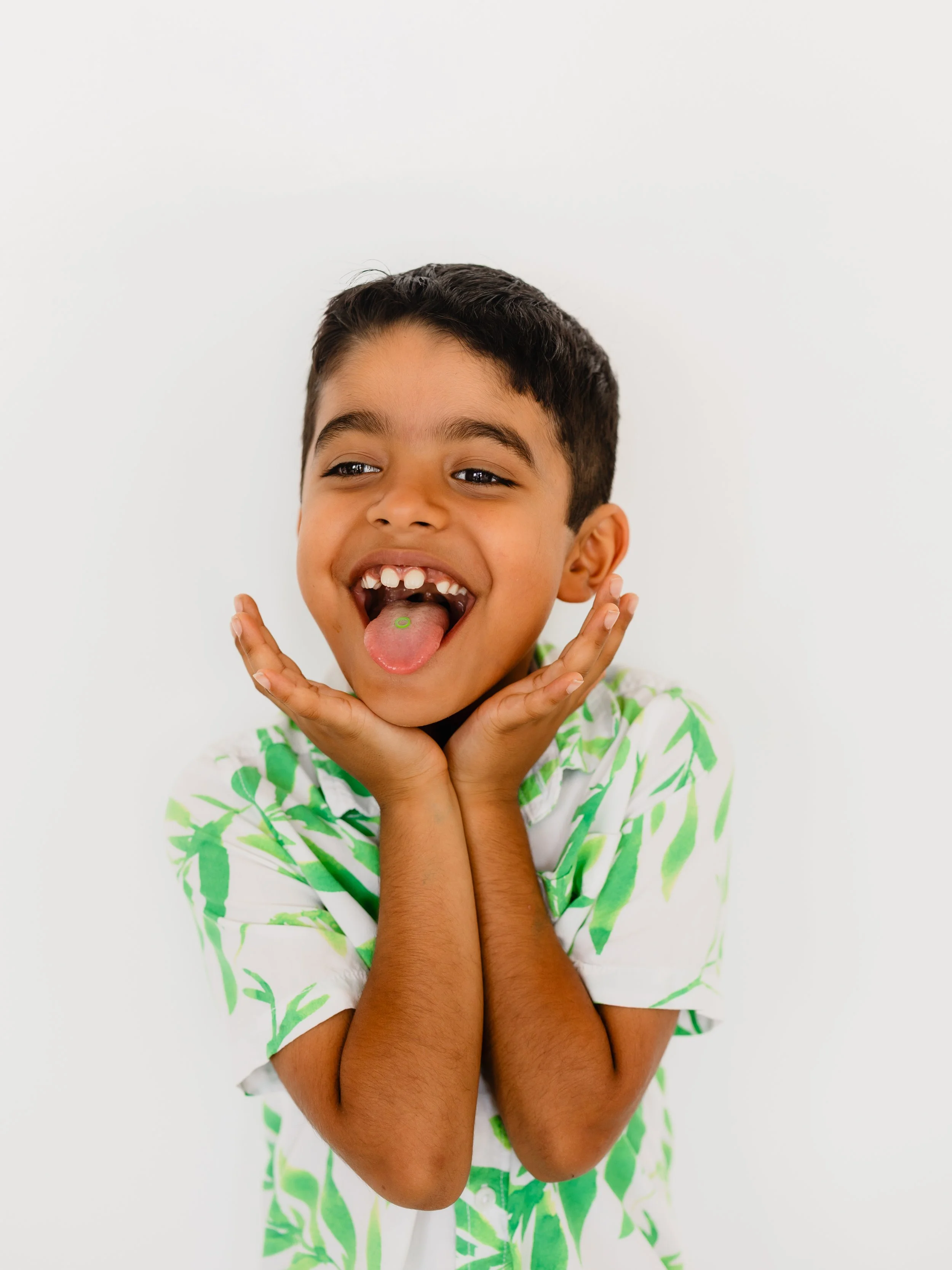 A young boy with dark hair smiling widely, sticking out his tongue, and touching his face with his hands. He's wearing a white shirt with green leafy patterns and has a dental band on his tongue.
