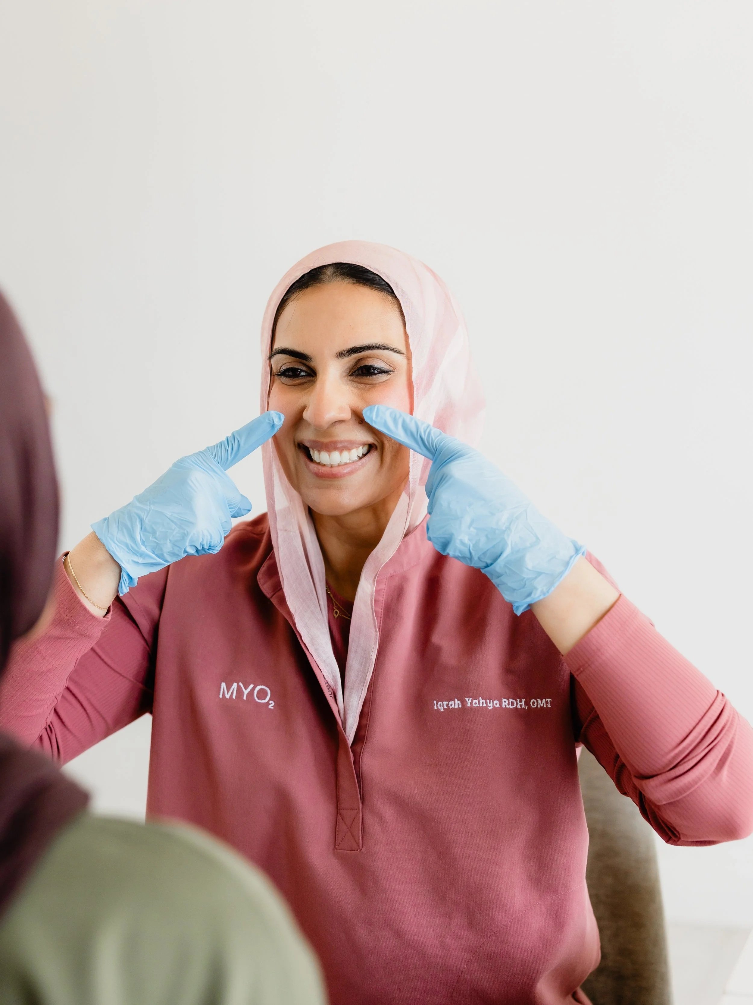 A smiling woman wearing a pink headscarf and a matching pink uniform with text, and gloves, having her cheeks touched by someone out of the frame.