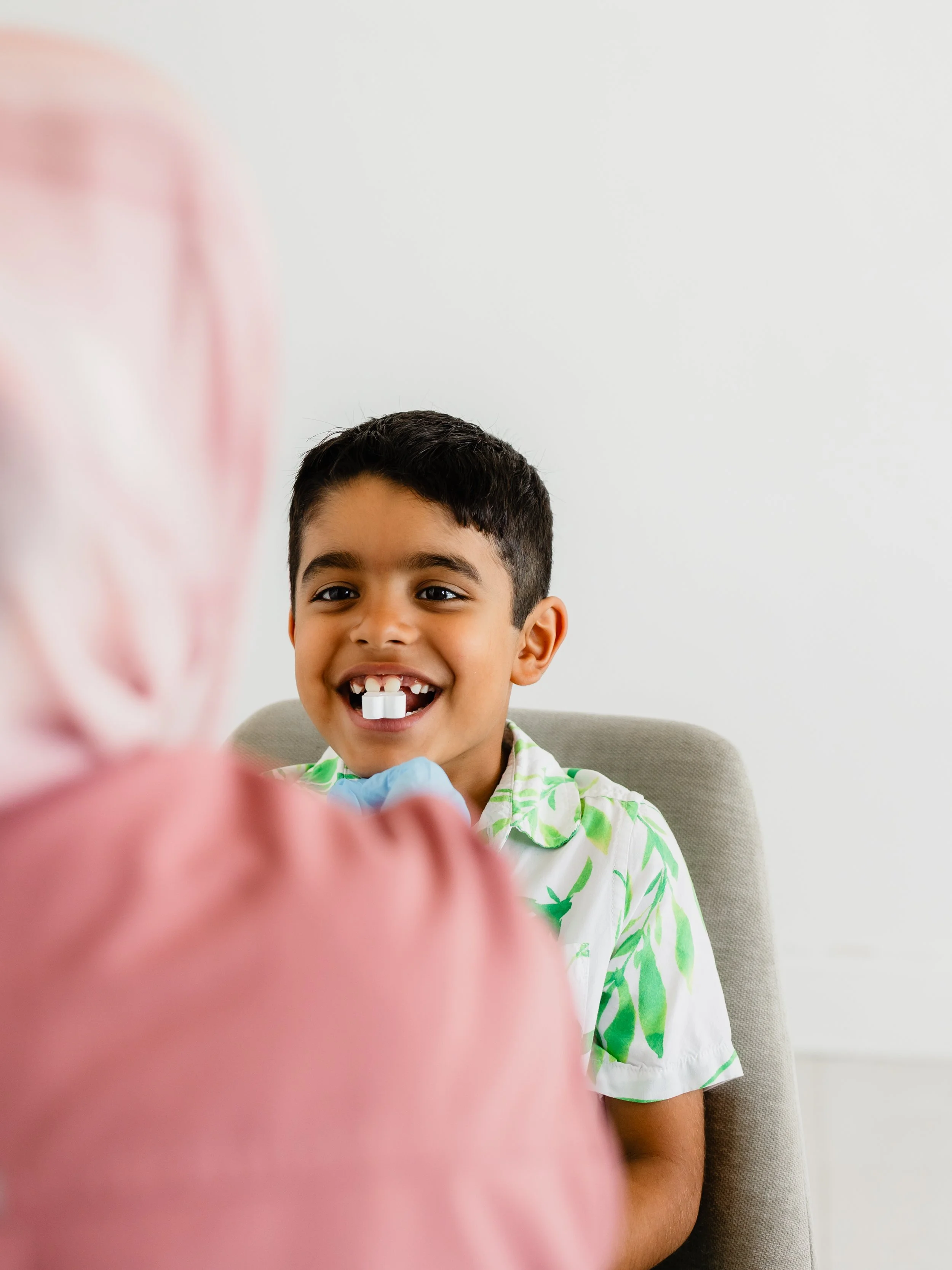 A young boy with short dark hair, smiling, showing missing teeth, sitting on a beige chair against a plain white wall, wearing a white shirt with green palm leaf pattern. The foreground is out of focus, showing a pink object.