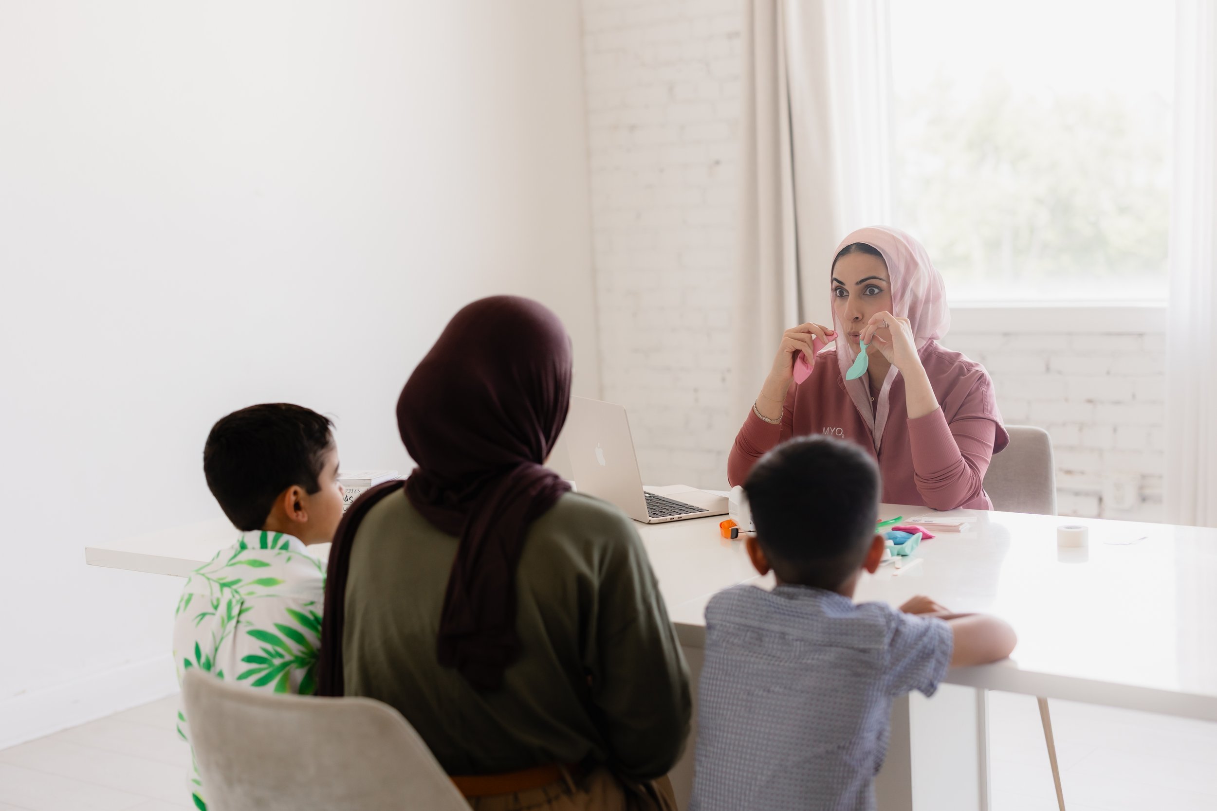A woman wearing a pink hoodie teaches a class of diverse children sitting at a white table in a bright room with white brick walls and large windows.