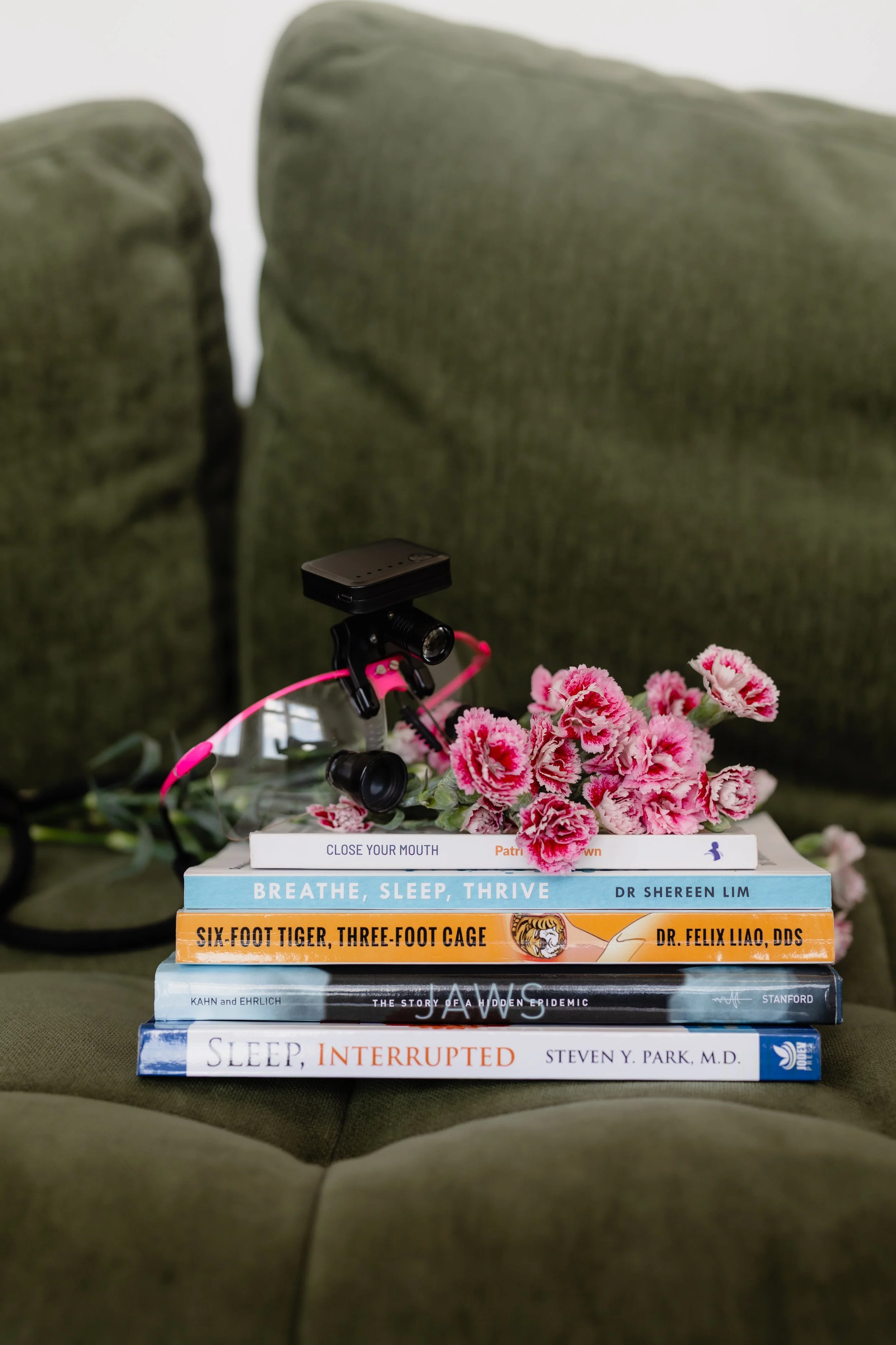 A stack of five books with pink flowers on top, placed on a green fabric surface. The books' titles include "Sleep, Interrupted" by Steven N. Park, M.D., "Sleep" by Kahn and Ehrlich, "Six-Foot Tiger, Three-Foot Cage" by Dr. Felix Liao, DDS, "Breathe, Sleep, Thrive" by Dr. Shereen Lim, and "Close Your Mouth" by Patrick McKeown.