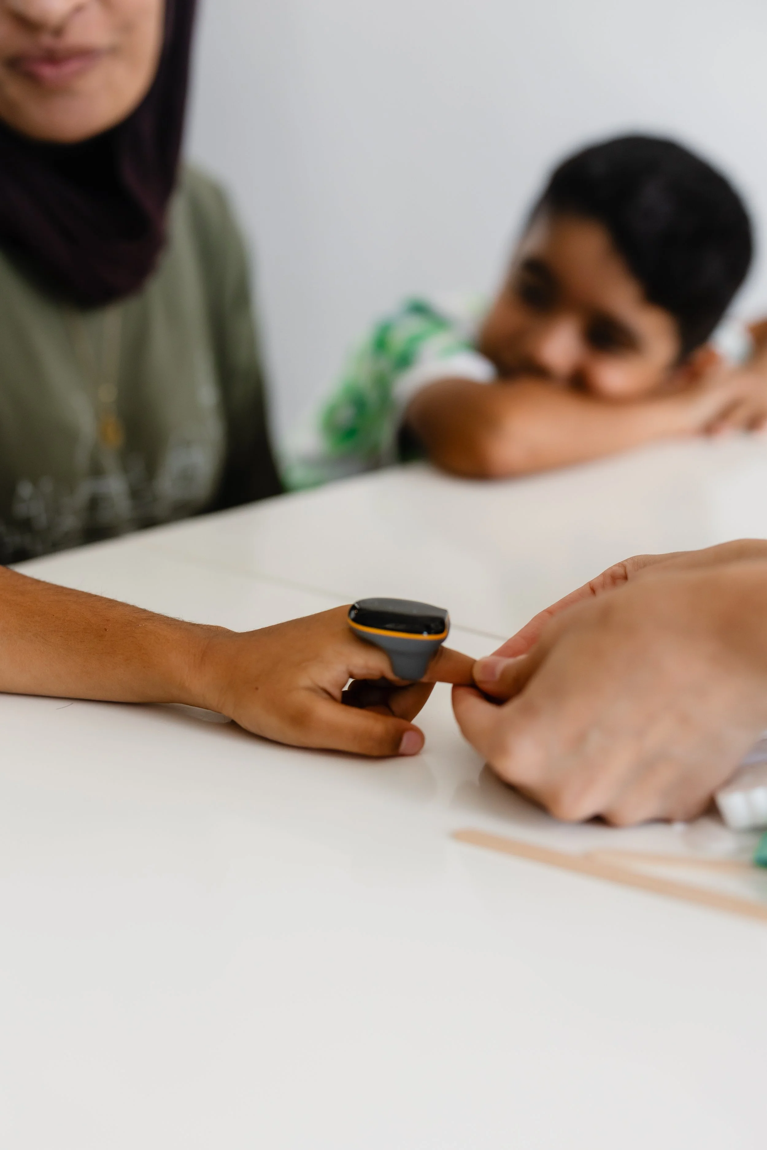 Close-up of an adult's hand holding a digital thermometer, with a child resting their head on their arm in the background, sitting at a white table.