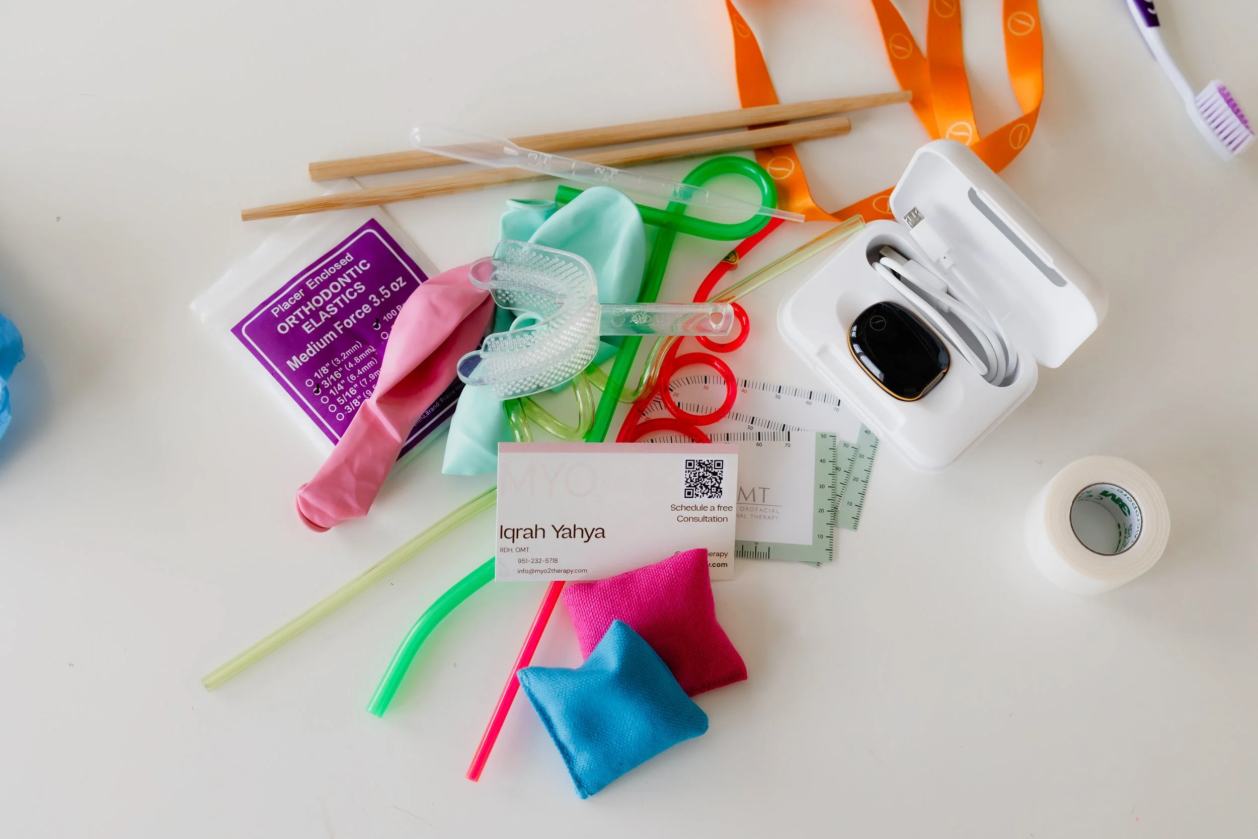 Medical supplies including colorful elastic bands, saliva sponges, a thermometer, business card, camouflage scrubs, and plastic pipettes scattered on a white surface.