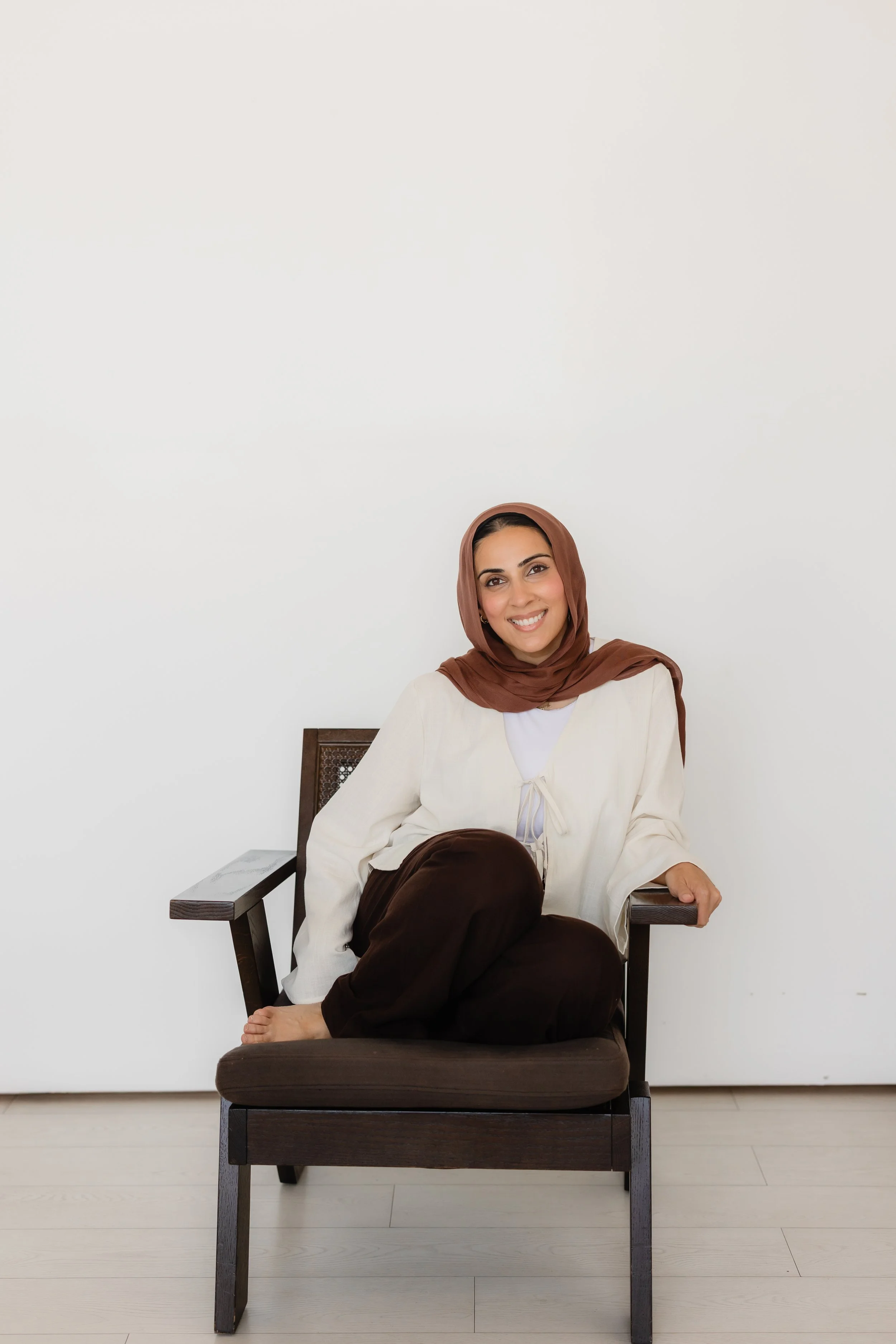 A woman wearing a brown headscarf and a cream-colored top sitting on a wooden chair with dark brown cushion, posed in front of a plain white wall.