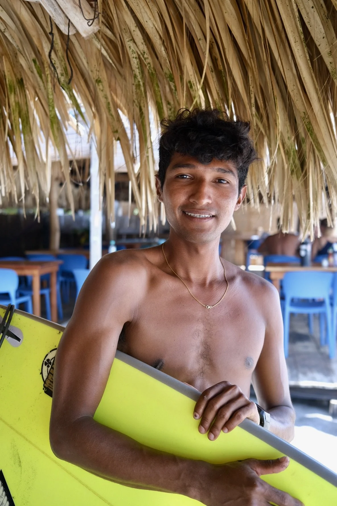 A shirtless man with short curly hair holding a yellow surfboard at a beachside hut.