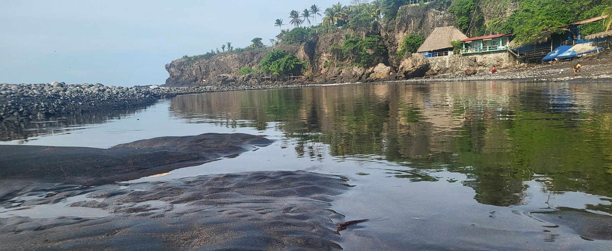 Calm ocean water reflecting a rocky shoreline with small boats, a cliff with green vegetation, and colorful beachside structures with palm trees in the background.