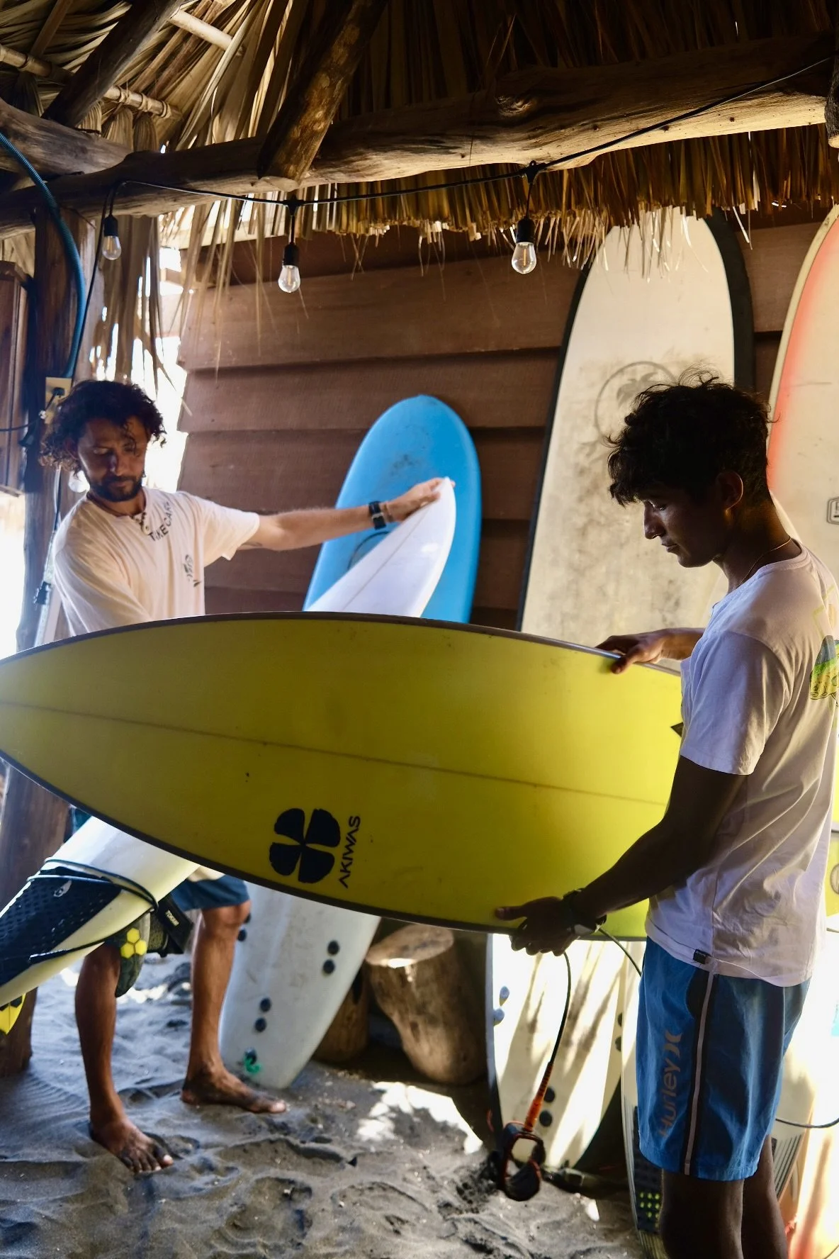 Two young men in a tropical, thatch-roof setting holding surfboards; one is yellow and the other is white with blue. There are additional surfboards leaning against the wall in the background. The scene appears to be at a surf shop or storage area with a sandy floor.