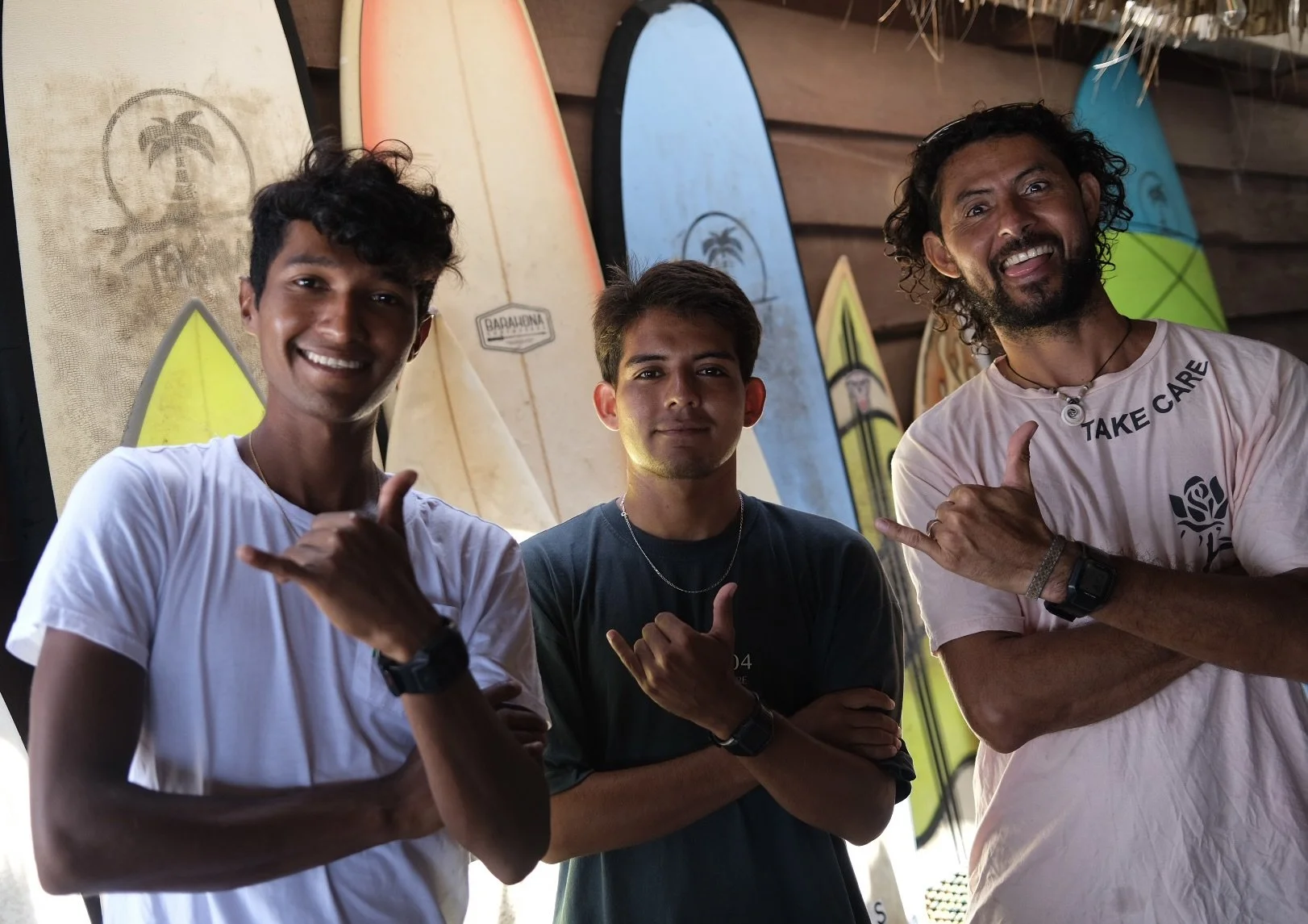 Three smiling men standing side by side, each making a shaka hand gesture, with surfboards displayed behind them.