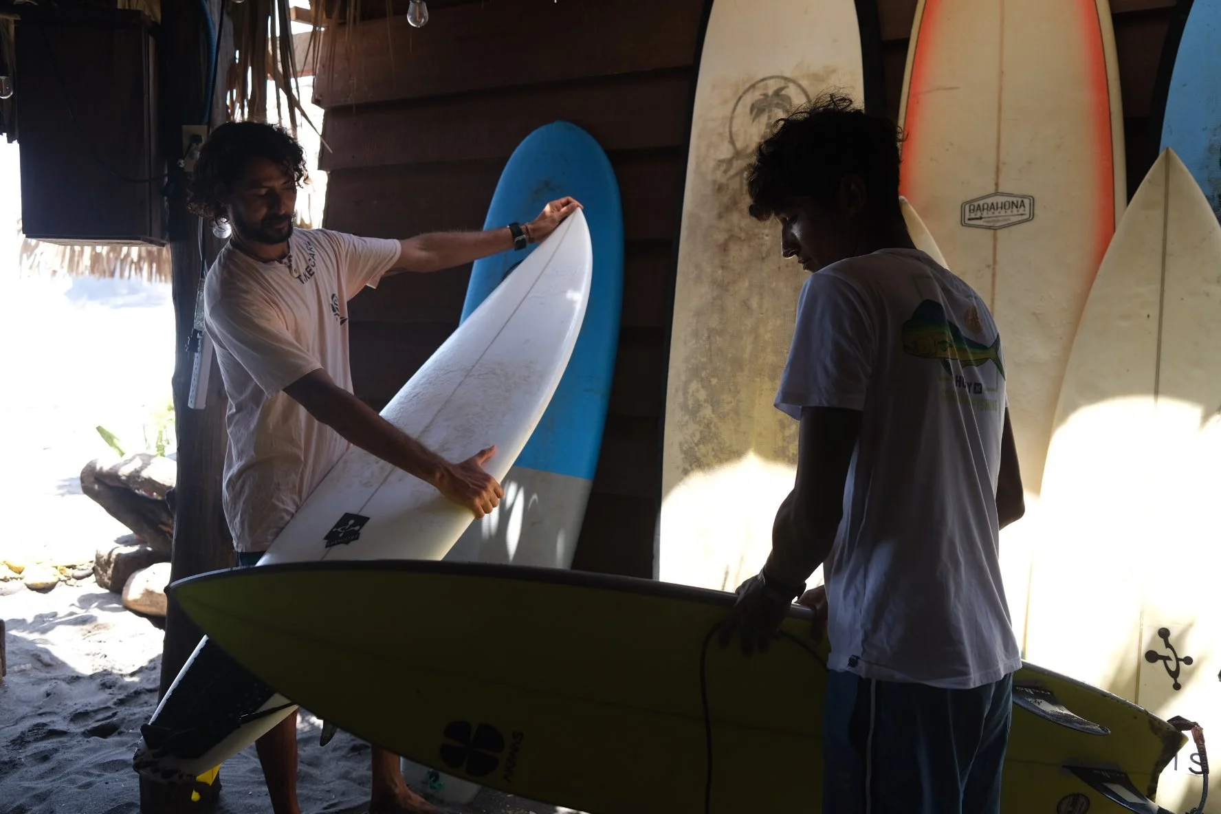Two men in a surf shop with surfboards, one showing a white surfboard to the other, surrounded by multiple surfboards, some with colorful designs, leaning against the wall.
