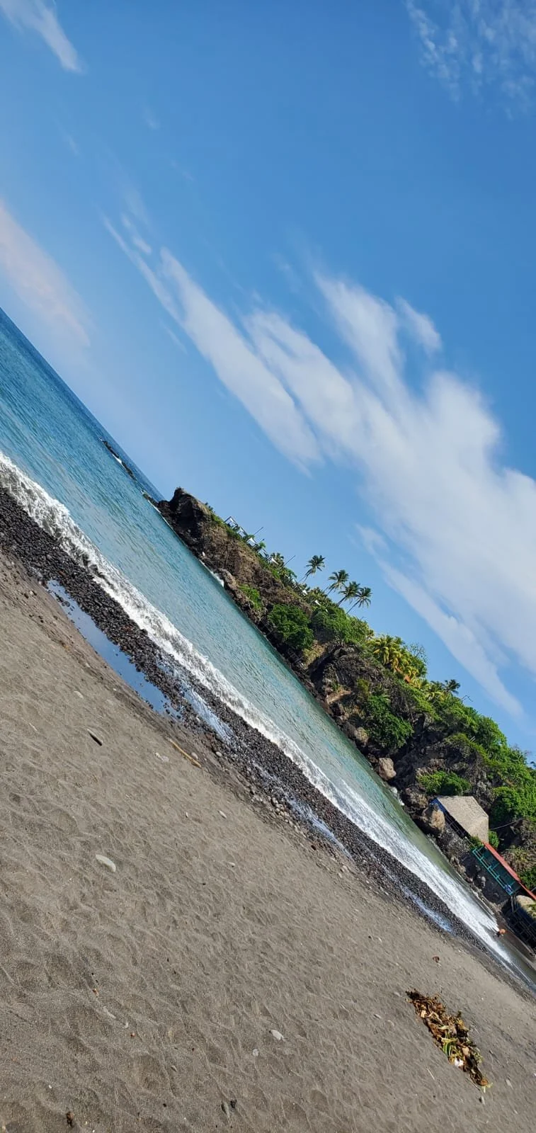 A beach with sandy shore, rocks, and clear blue water under a partly cloudy sky, with palm trees and greenery on a rocky landmass in the background.