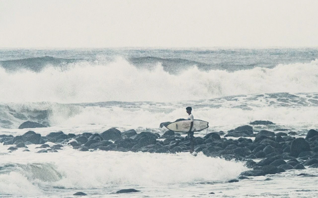 A person standing on rocks by the ocean holding a surfboard, with large waves in the background.