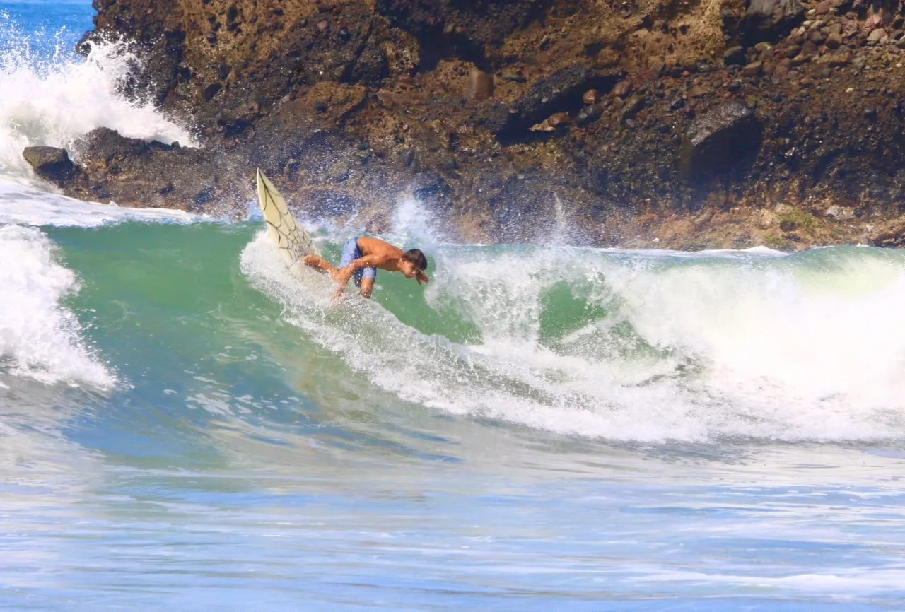 A surfer dressed in blue shorts riding a wave near rocky shoreline.