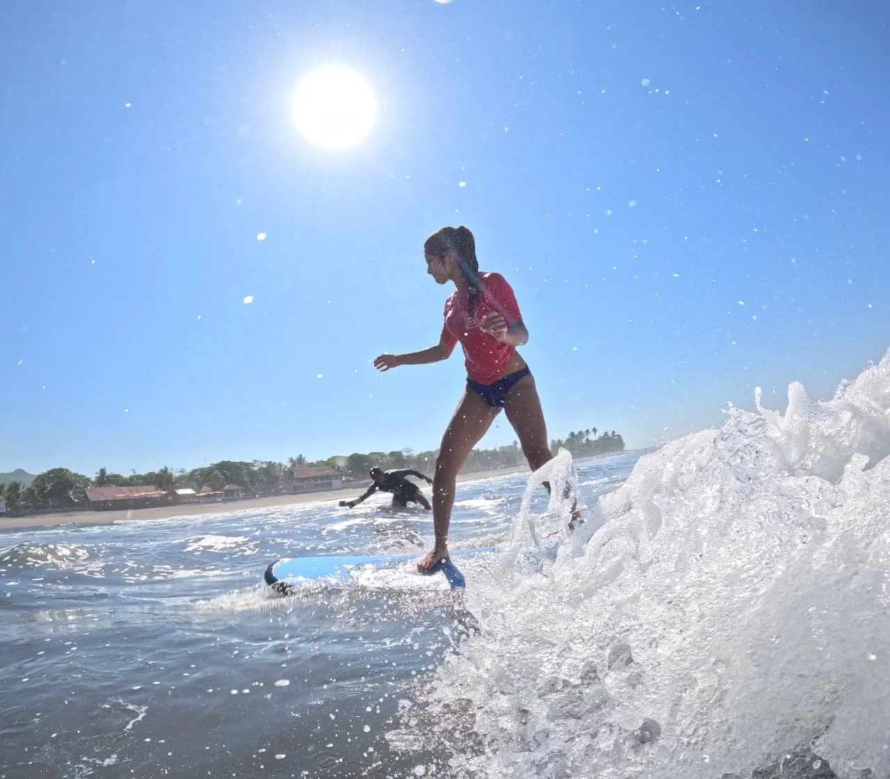 A woman in a red shirt and black shorts riding a surfboard on a sunny beach, with a wave splashing around her and another person in the water in the background.