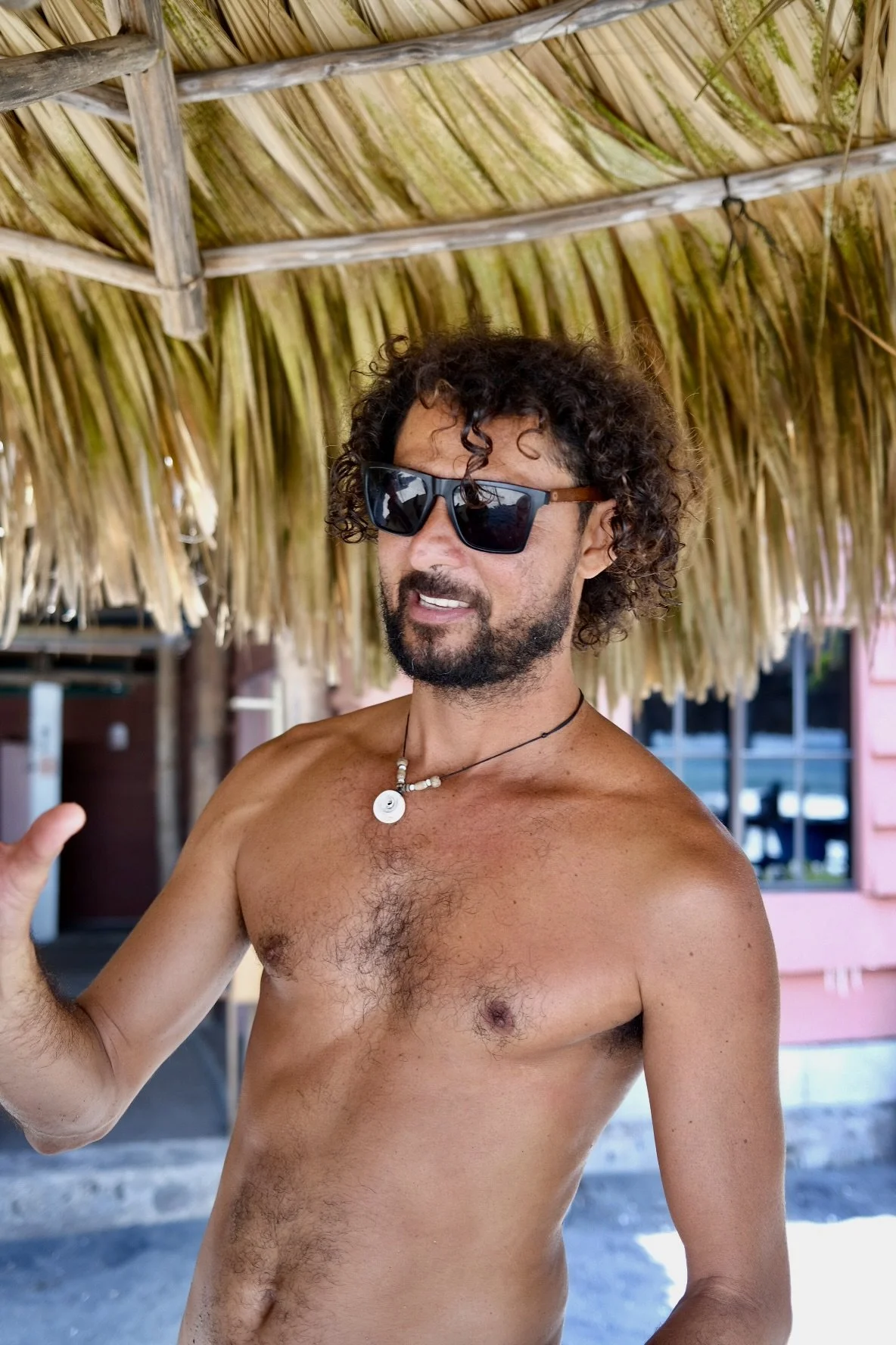 A shirtless man with curly hair and sunglasses, under a thatched roof, at a tropical beach or resort.