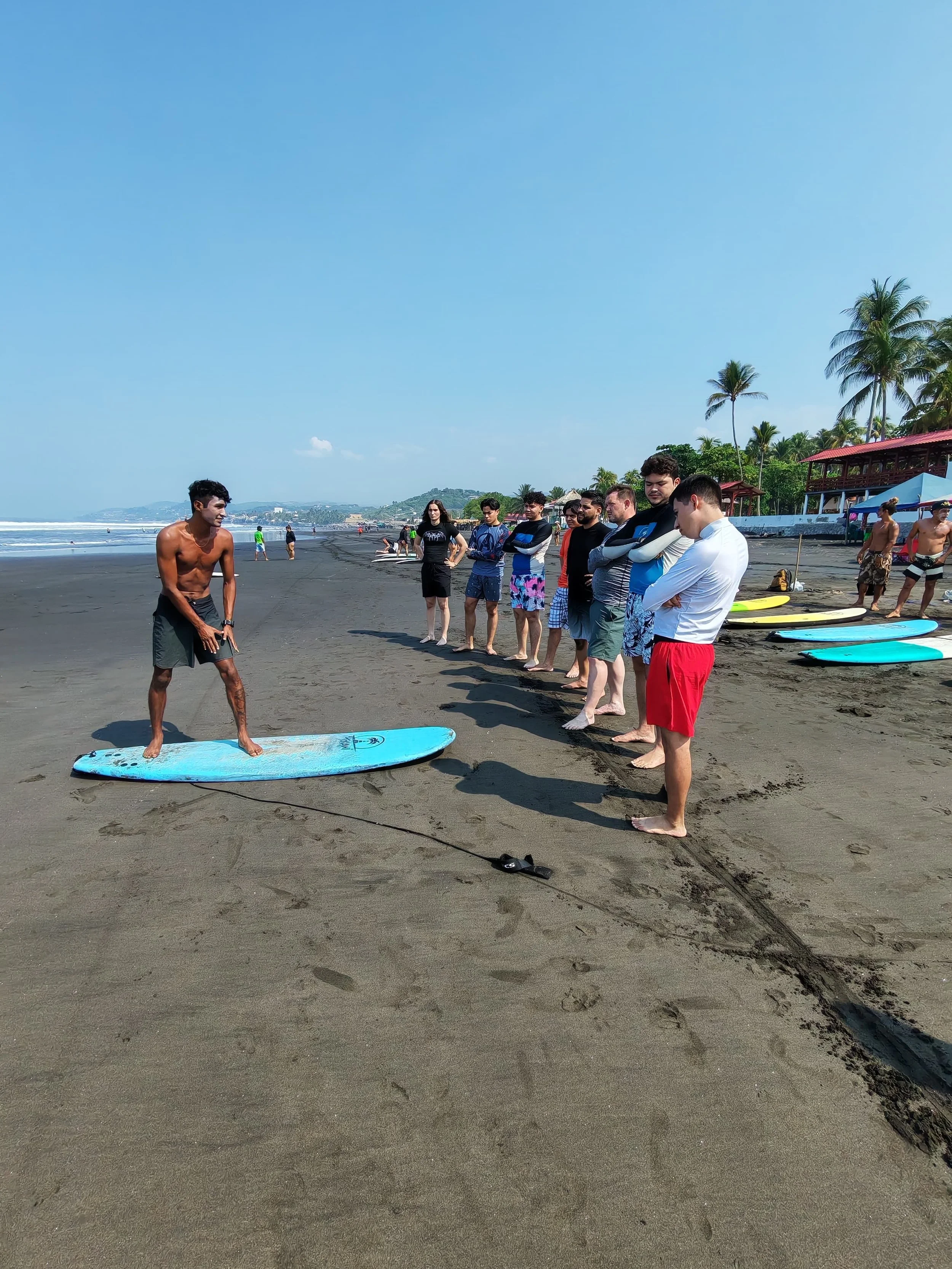 A group of people on a beach, some standing in a line and a shirtless man on a surfboard.