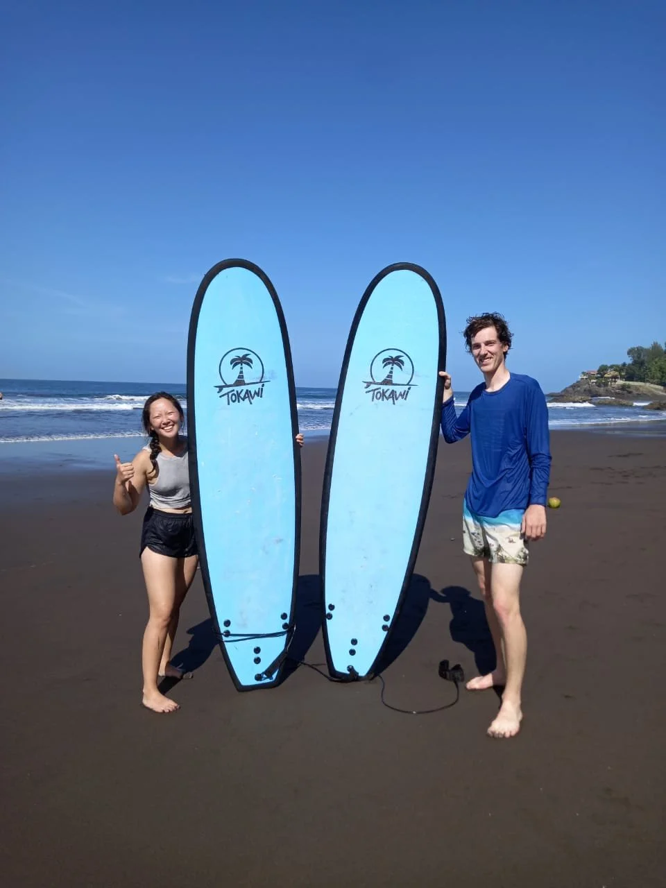 Two people holding large blue surfboards with the word 'Tokawi' and a palm tree logo, standing on a sandy beach with the ocean and clear blue sky in the background.