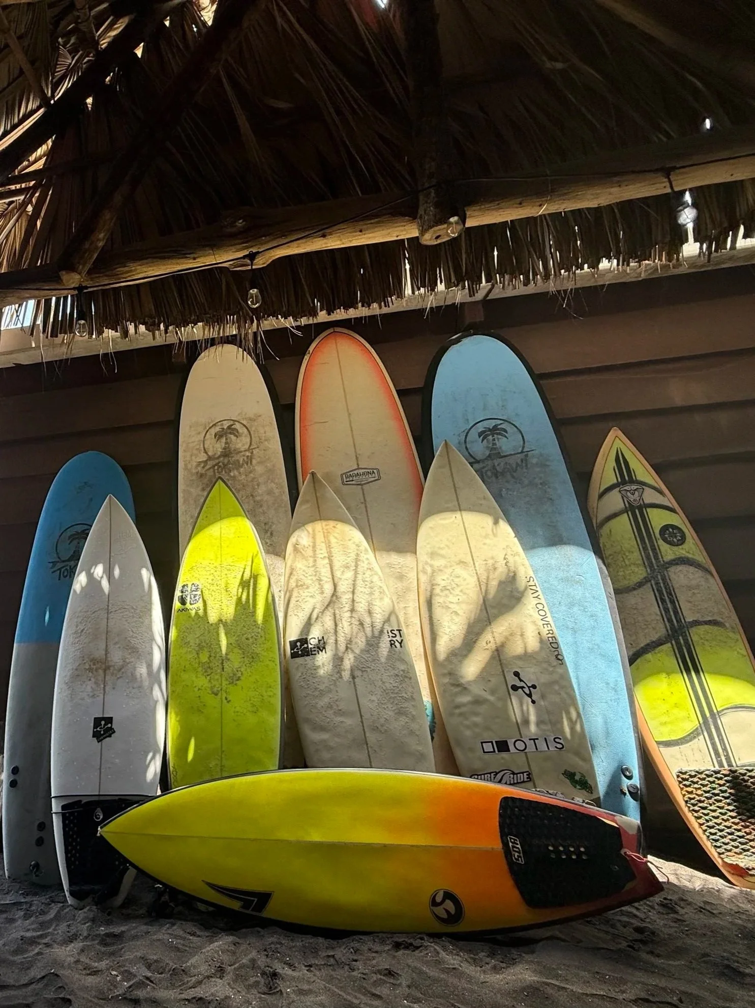 A row of colorful surfboards leaning against a wall under a thatched roof on a sandy beach.