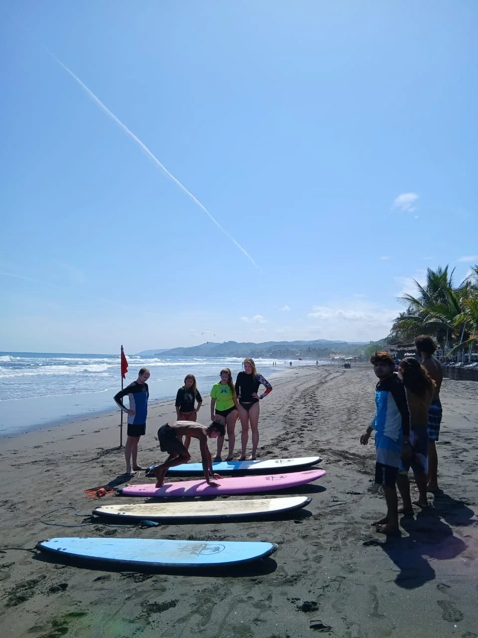 Group of people gathered on a sandy beach with paddleboards, some standing and one crouching on a pink paddleboard, with the ocean, palm trees, and a clear blue sky in the background.
