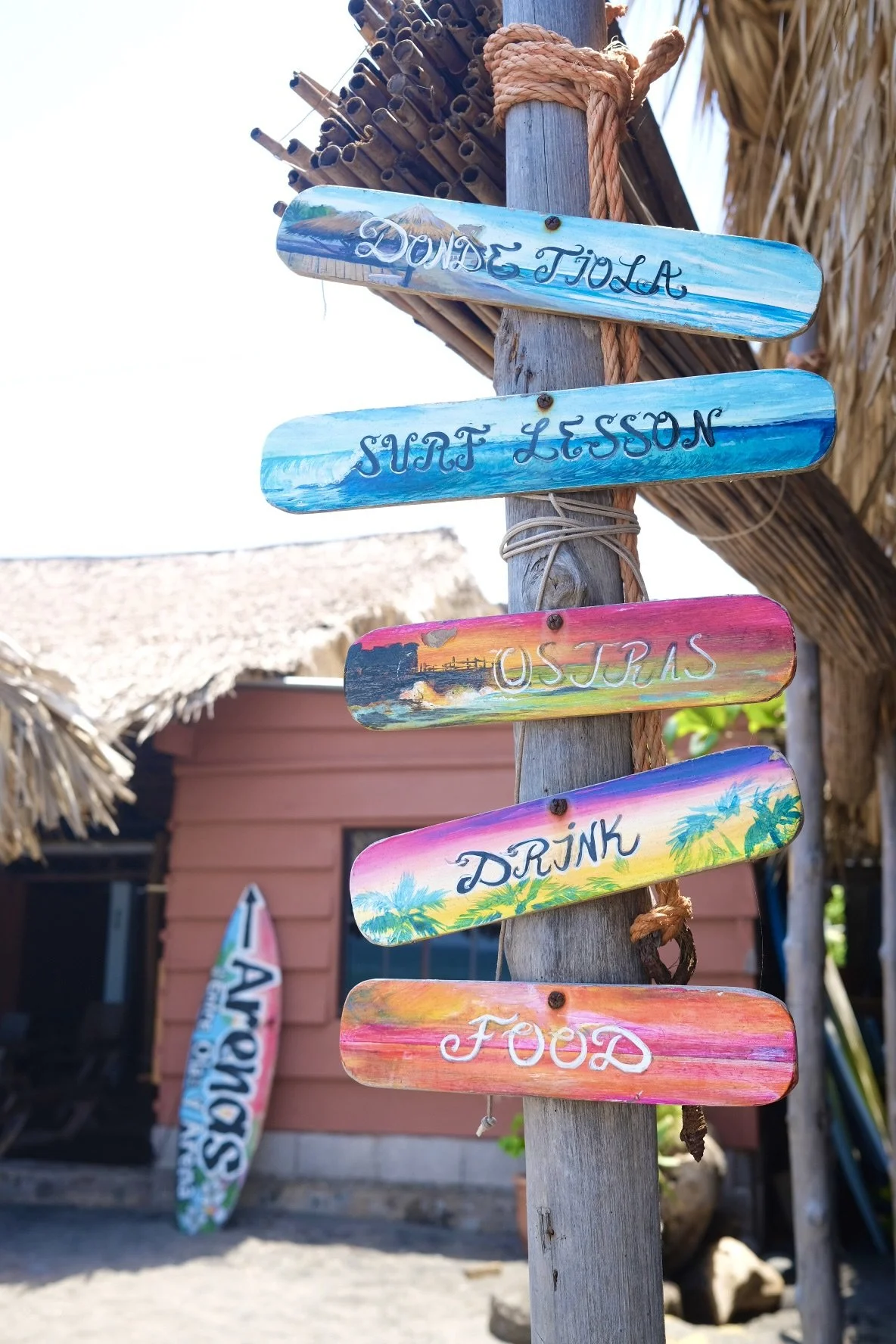 Colorful wooden signs with handwritten text pointing to different beach activities, including 'Dunce Tola', 'Surf Lesson', 'Ostias', 'Drink', and 'Food', attached to a weathered wooden post with a rope, with a thatched-roof building and a surfboard in the background on a beach.