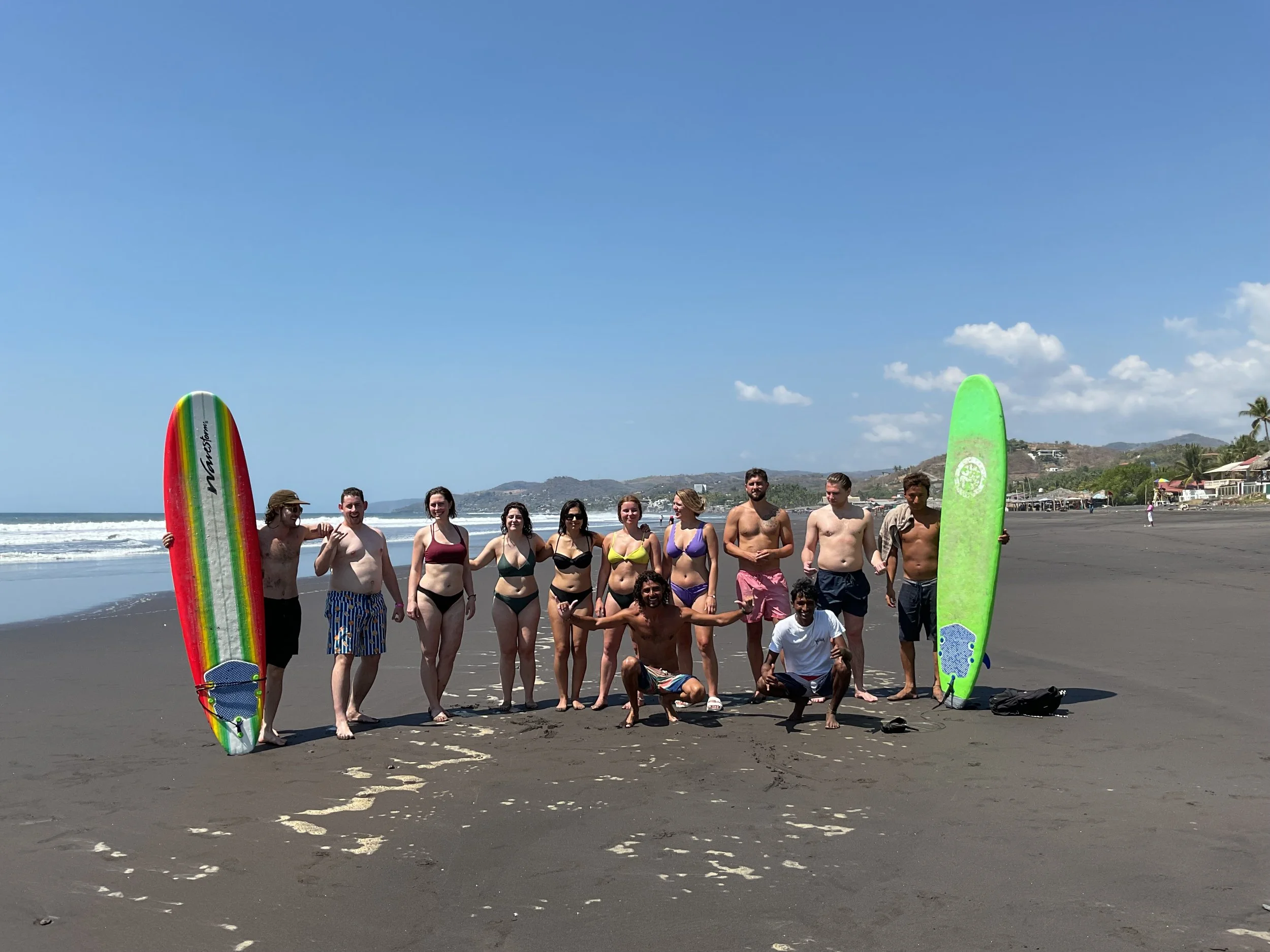 A group of people standing on a beach with surfboards, some wearing swimsuits and sunglasses, with a cloudy blue sky and mountains in the background.