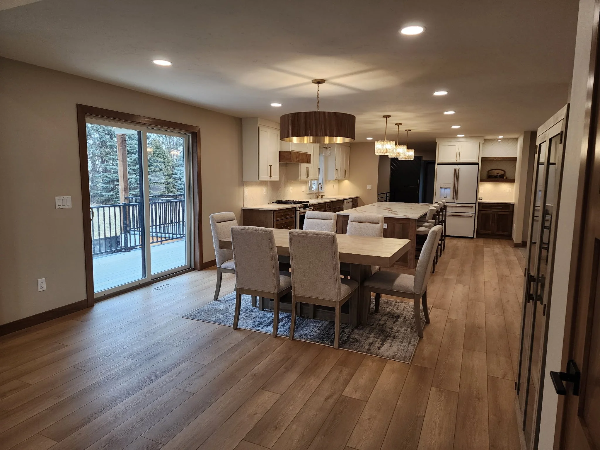 Open-concept kitchen and dining area with wood flooring, large window, sliding glass door leading outside, white cabinets, kitchen island with barstools, and modern pendant lighting.