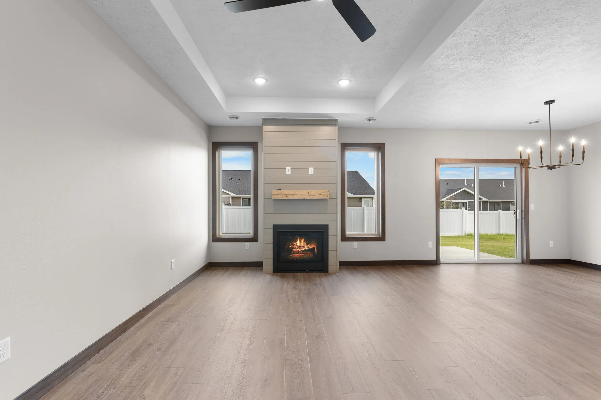 Empty living room with light-colored hardwood floors, white walls, a fireplace, two narrow windows, and sliding glass door leading outside, with a ceiling fan and chandelier.