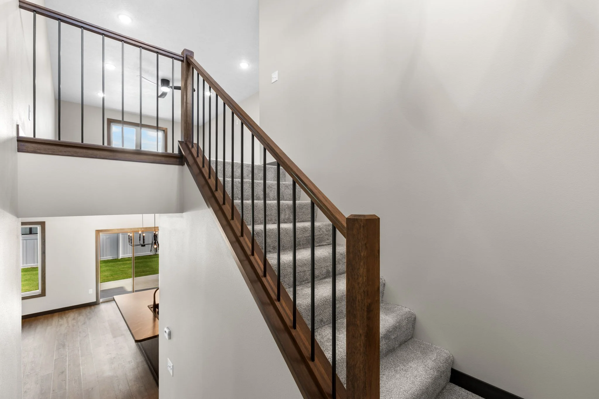 Interior view of a staircase with grey carpeted steps, black metal spindles, and a wooden handrail, leading to a second floor in a modern home with white walls and ceiling lights.
