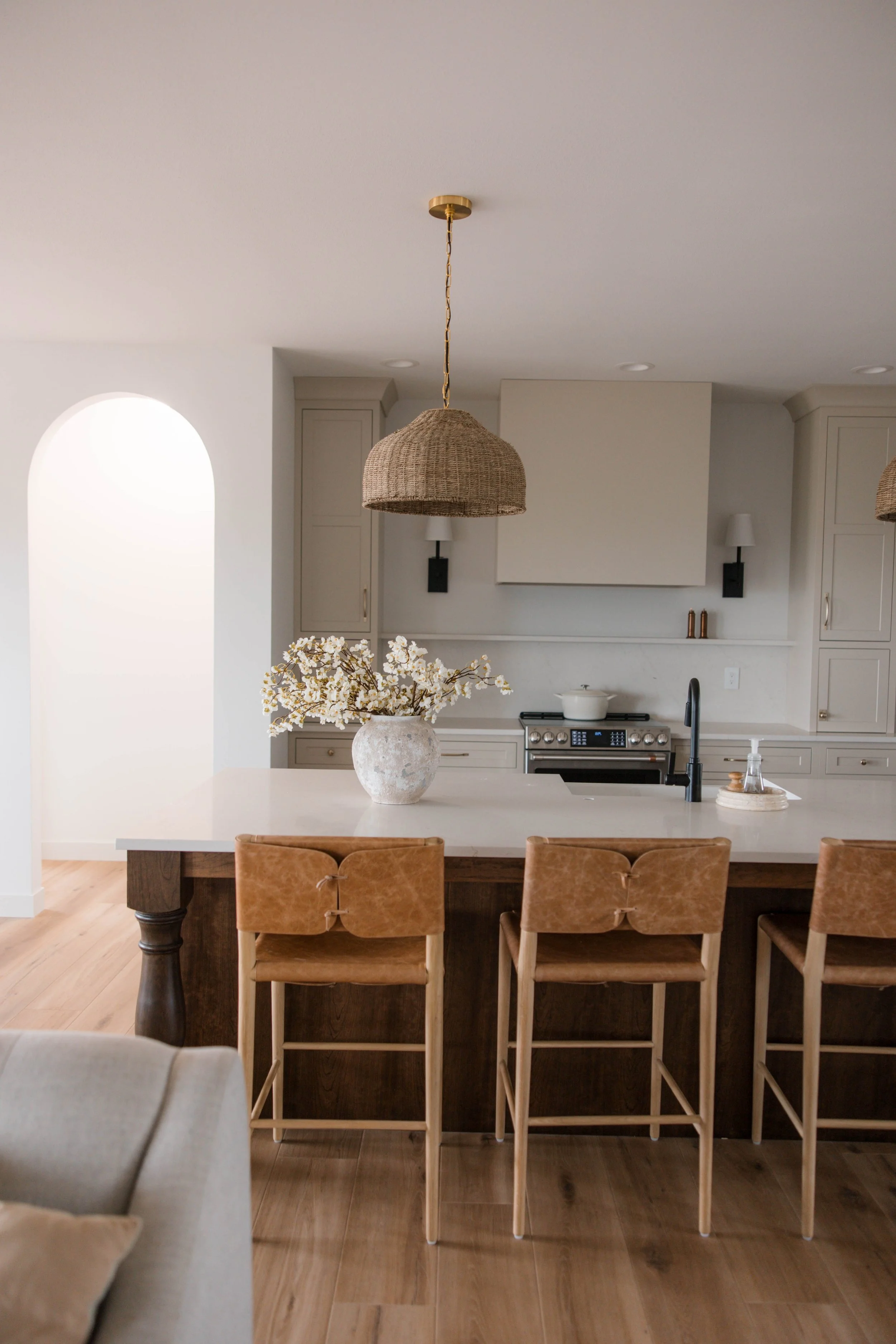 Modern kitchen with white cabinets, stainless steel stove, and a large island. Four light brown bar stools are positioned at the island. A large vase with white flowers decorates the island. Woven pendant light hangs above the island. The background 