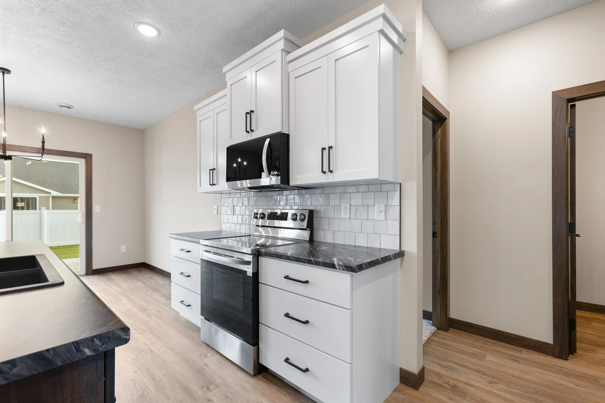 Modern kitchen with white cabinets, black handles, stainless-steel microwave, and stove, with a gray tile backsplash and wooden flooring.