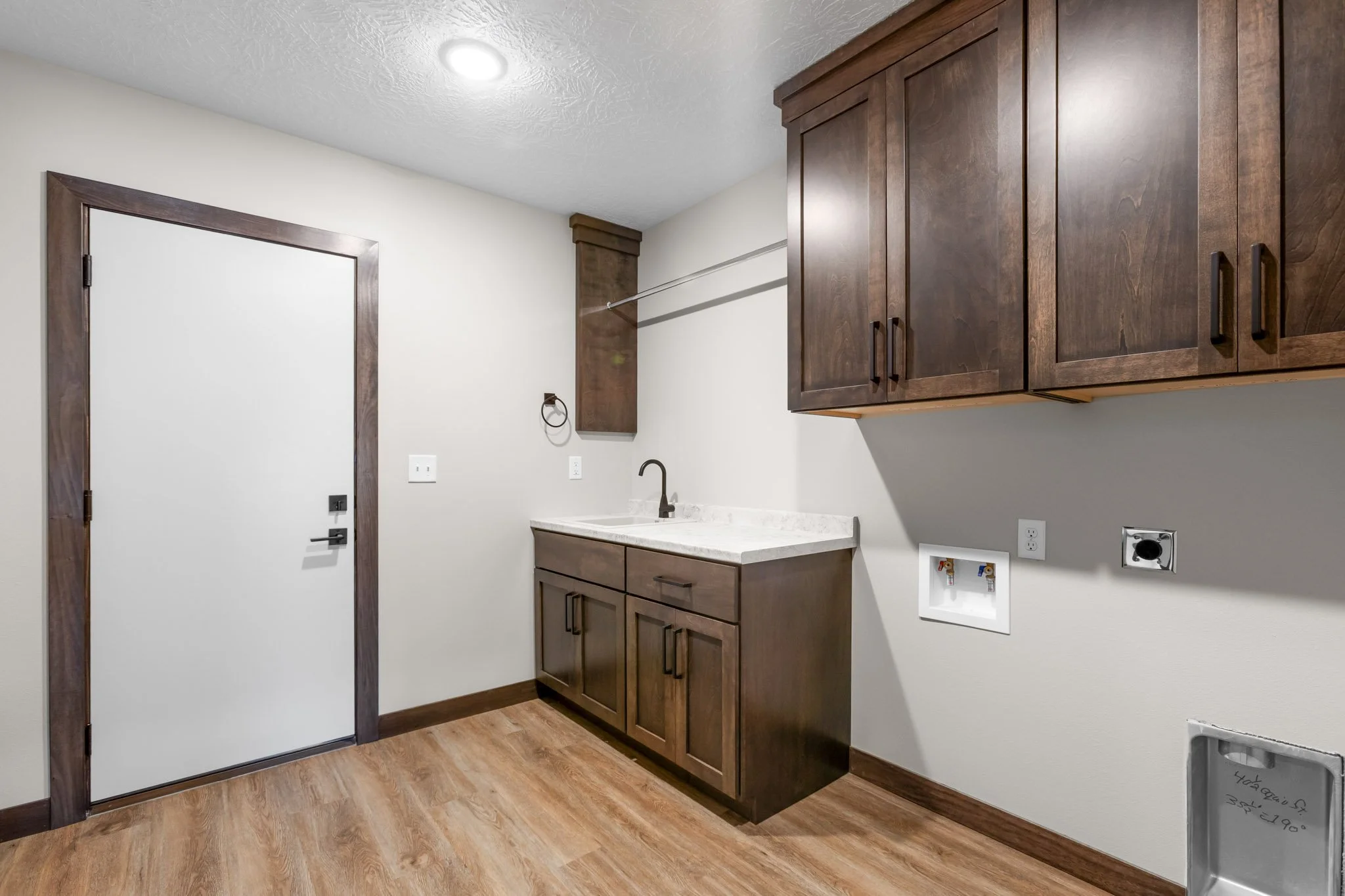 Interior view of a laundry room with wooden cabinets, a countertop with a sink, a ceiling light, and an opening for appliances, with wood-style flooring and a door frame.