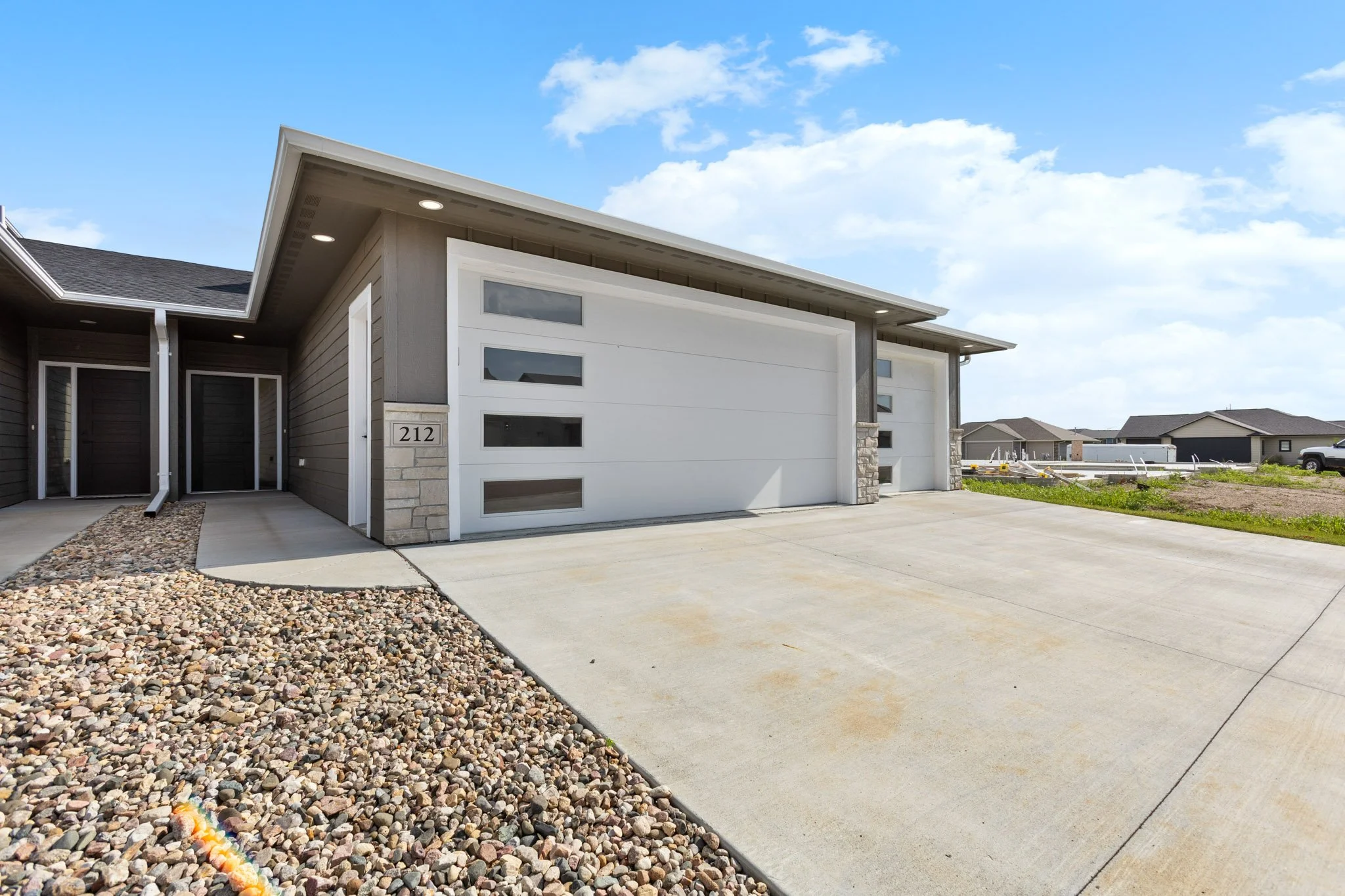 Modern house with a concrete driveway and a rock bed in front, under a bright blue sky with a few clouds.