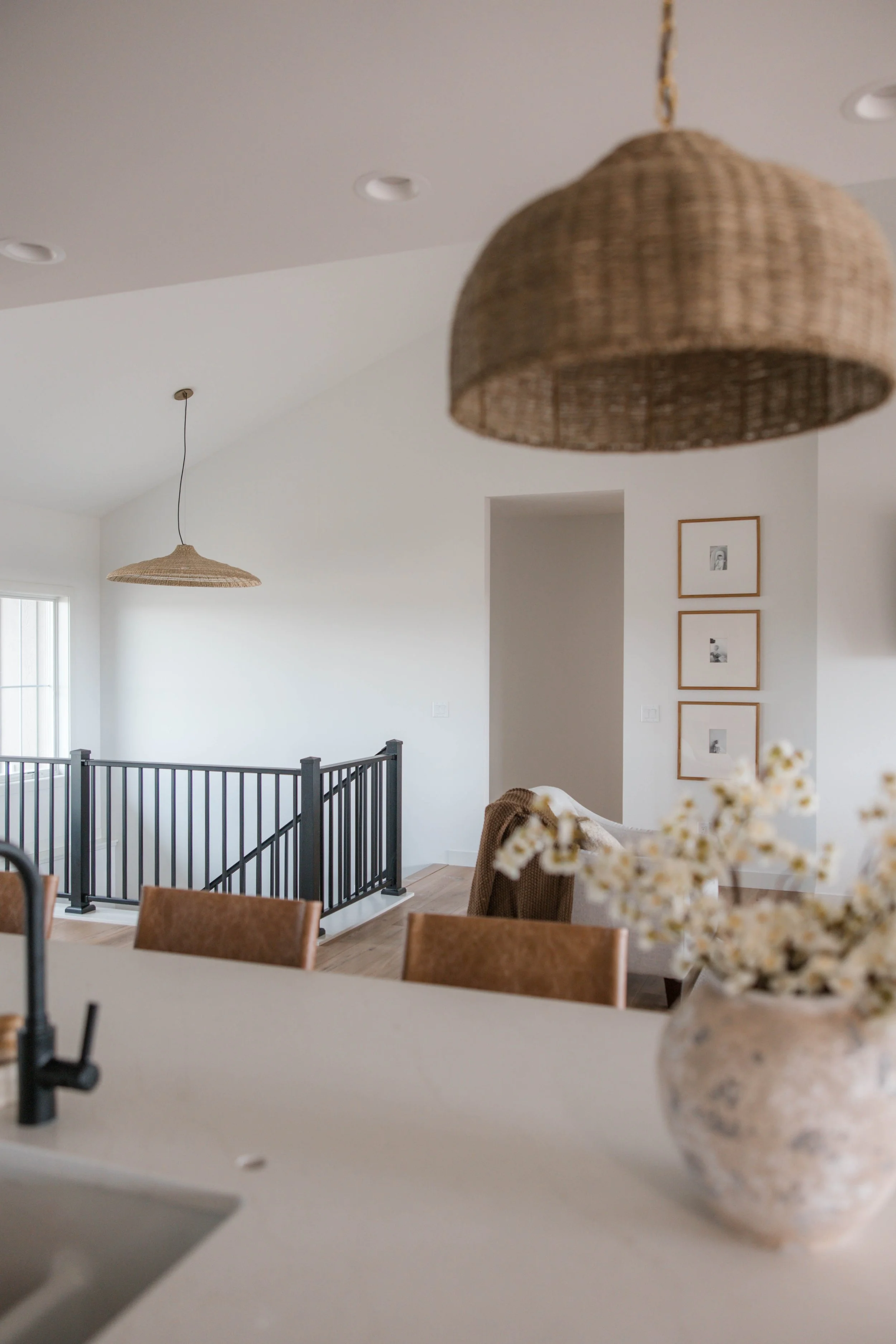 Living room with a hanging woven light fixture, white walls, framed black and white pictures, and a black railing around a stairway or balcony.