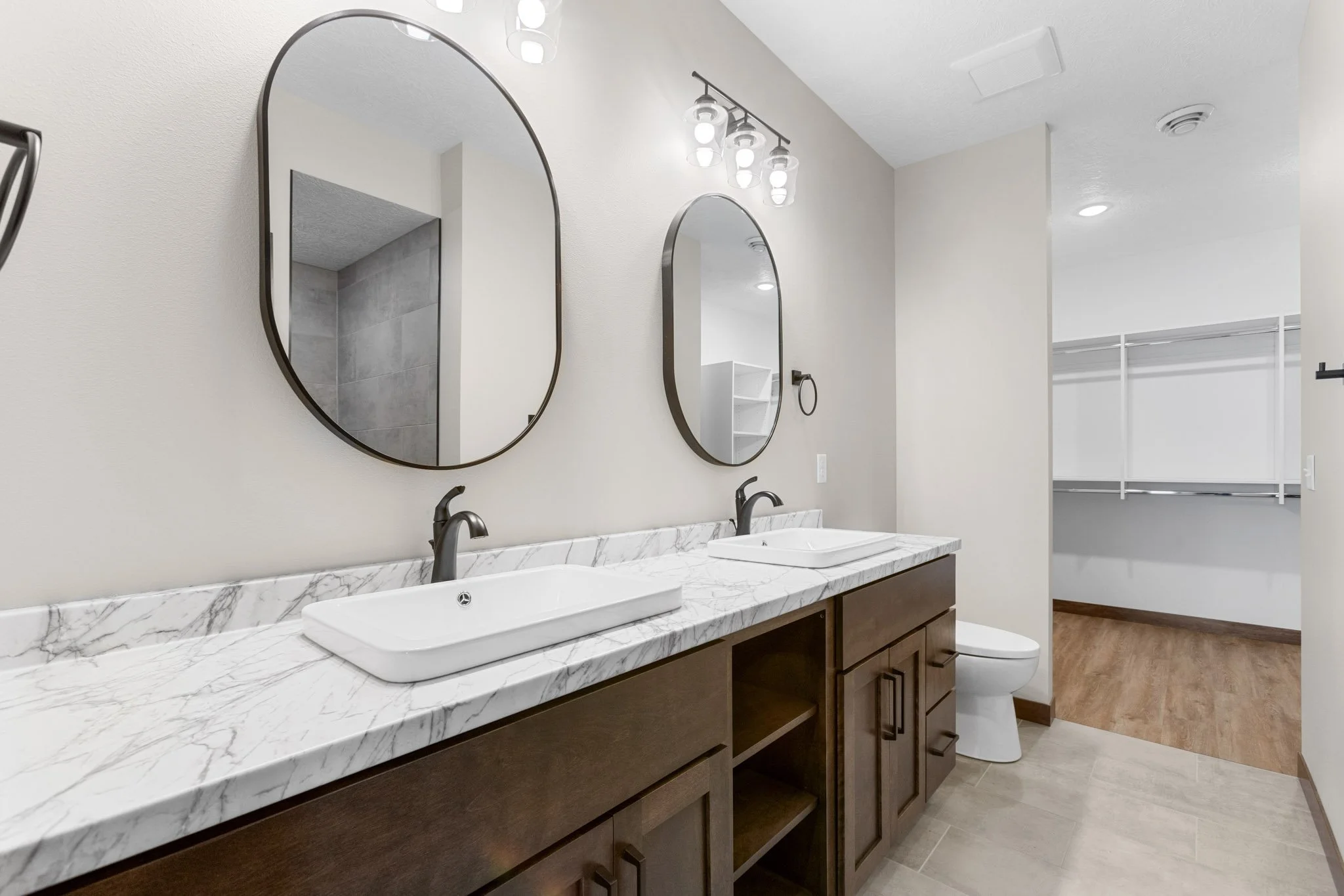 A modern bathroom with two rectangular white sinks on a marble countertop, two round mirrors with black frames, a dark wood vanity, a wall-mounted faucet, and a wall-mounted light fixture with multiple bulbs.