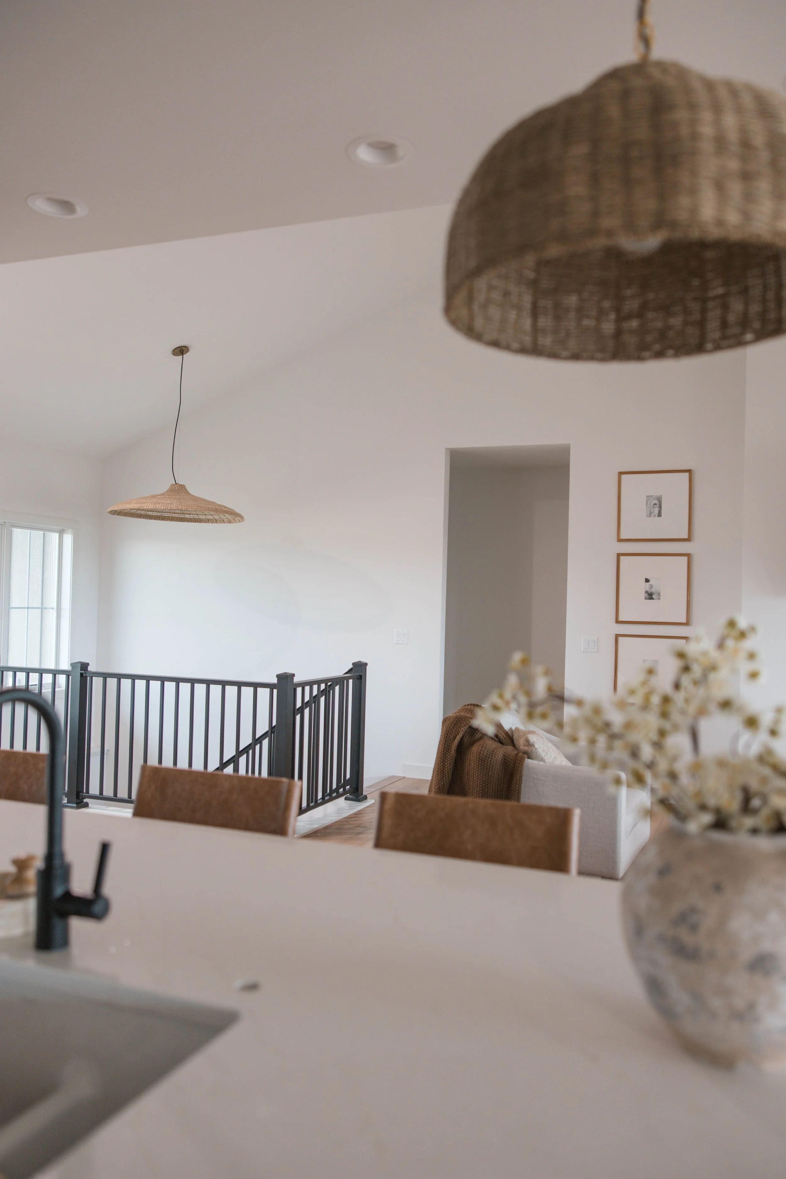 A modern, minimalistic interior with white walls, two woven pendant lights hanging from the ceiling, framed black and white photographs on the wall, a beige sofa with a brown throw, a black metal staircase railing, and a cozy dining area with wooden 