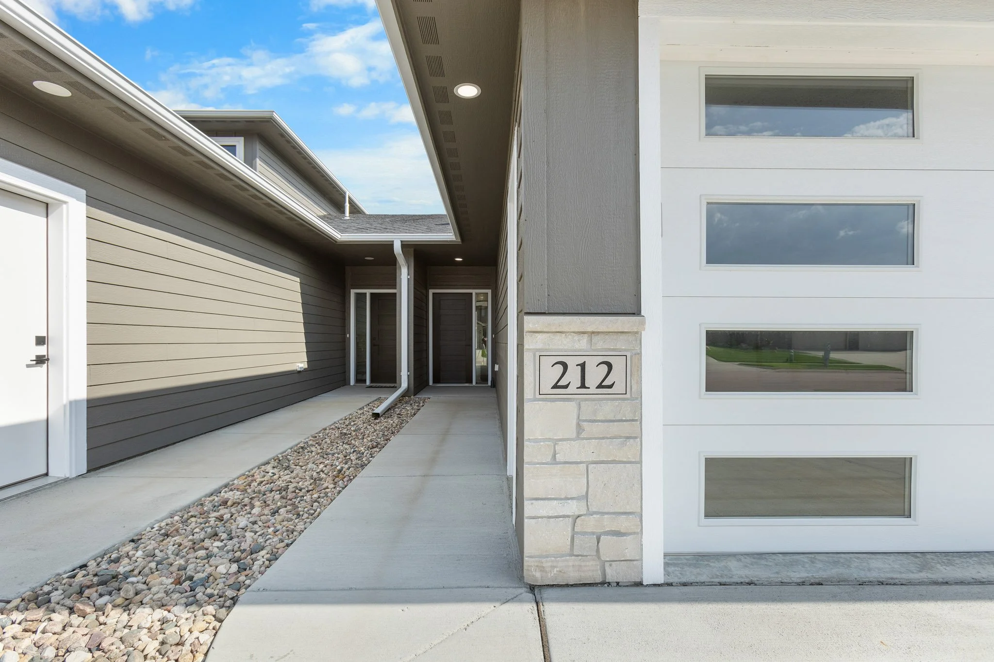 Exterior view of a modern house with a driveway, beige and white siding, large windows reflecting a cloudy sky, and house number 212 displayed on a stone pillar.