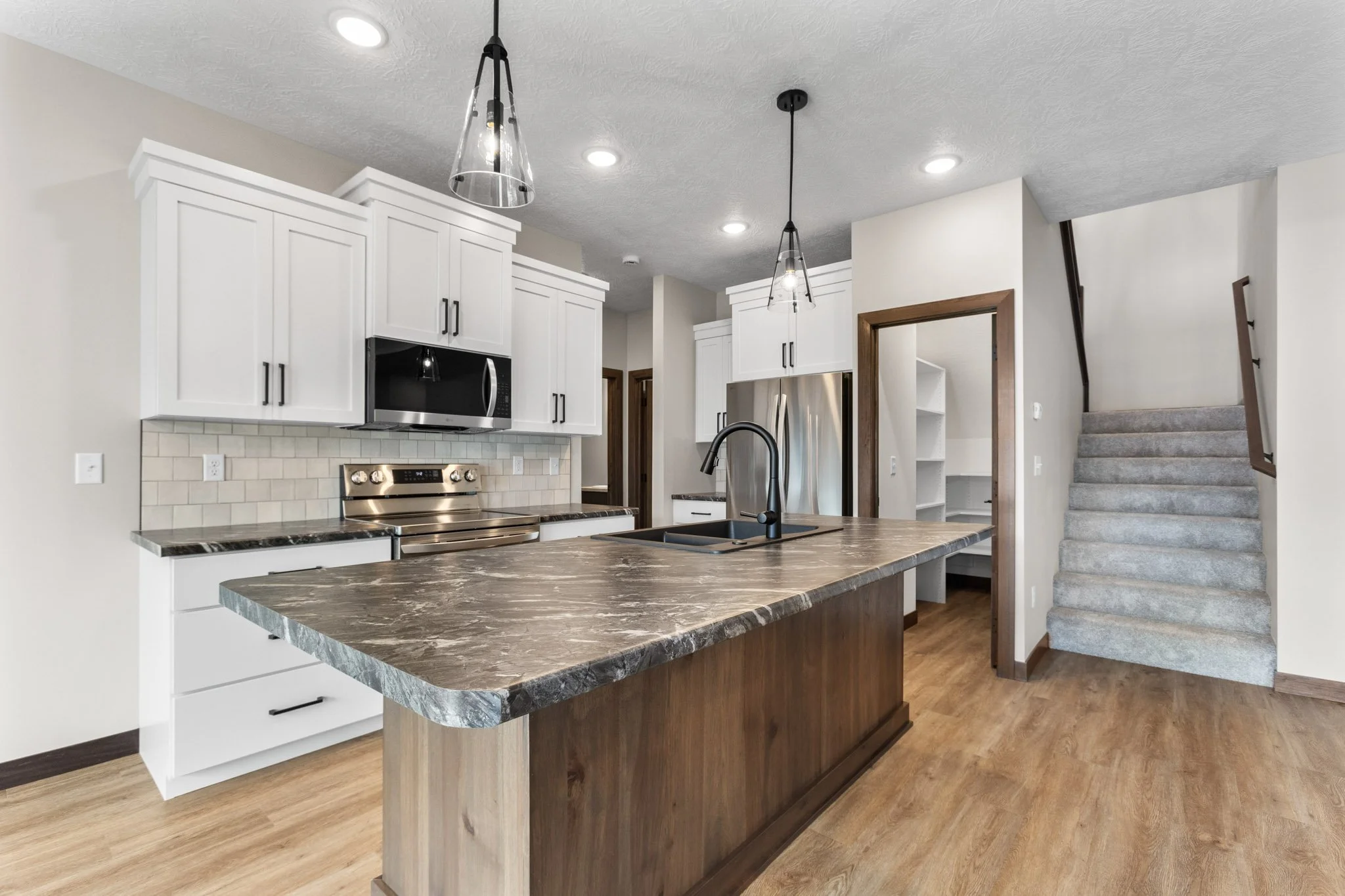 Modern kitchen with white cabinets, a black and gray marble kitchen island, black faucet, stainless steel appliances including a microwave and stove, and a staircase leading upstairs.