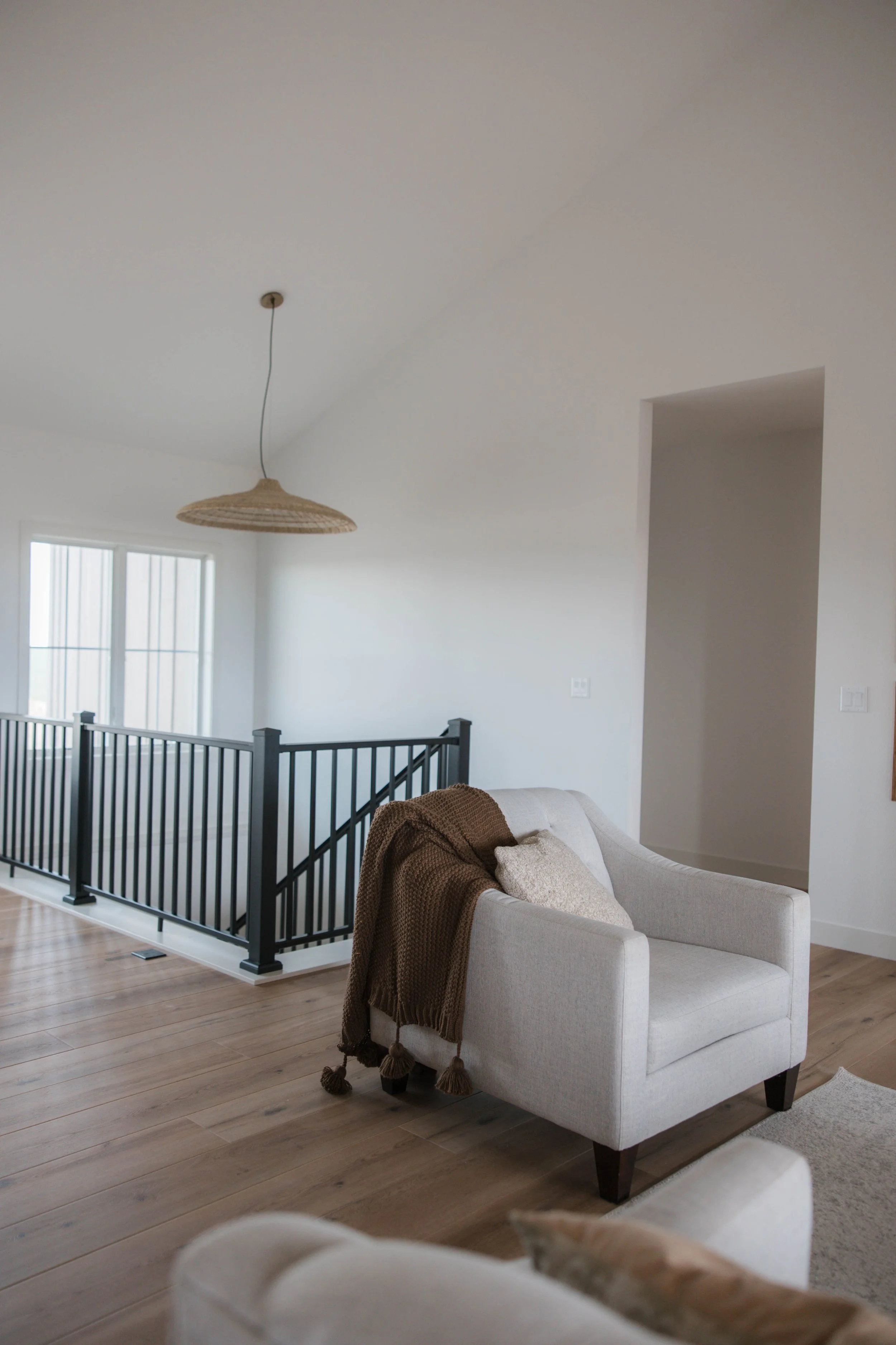 Living room with white walls, hardwood floors, beige armchair with brown throw blanket, black metal railing, window, and hanging woven light fixture.