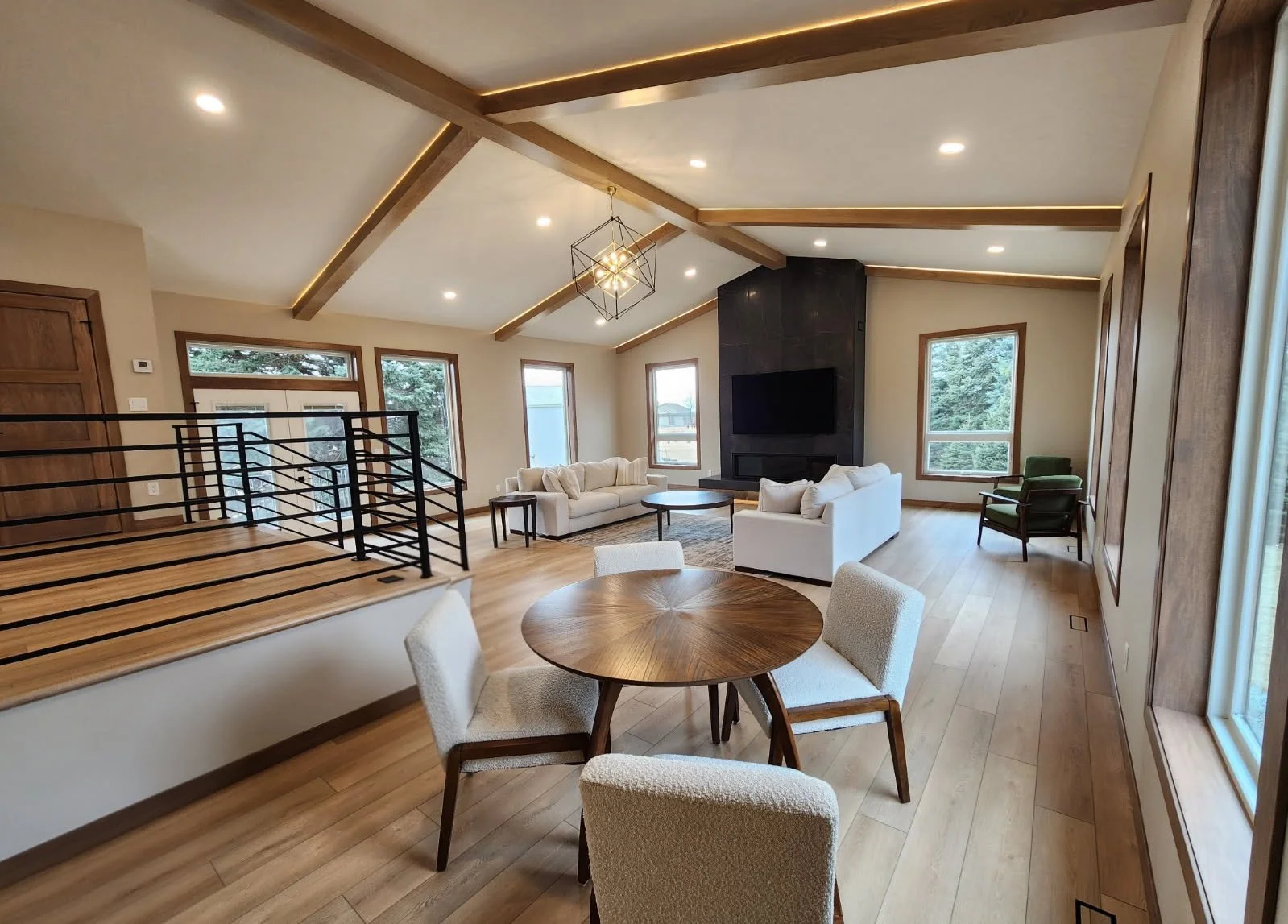 Open-concept living room with wood floors, a round dining table with four chairs, white sofas arranged around a black fireplace with a TV above, and multiple large windows providing natural light.