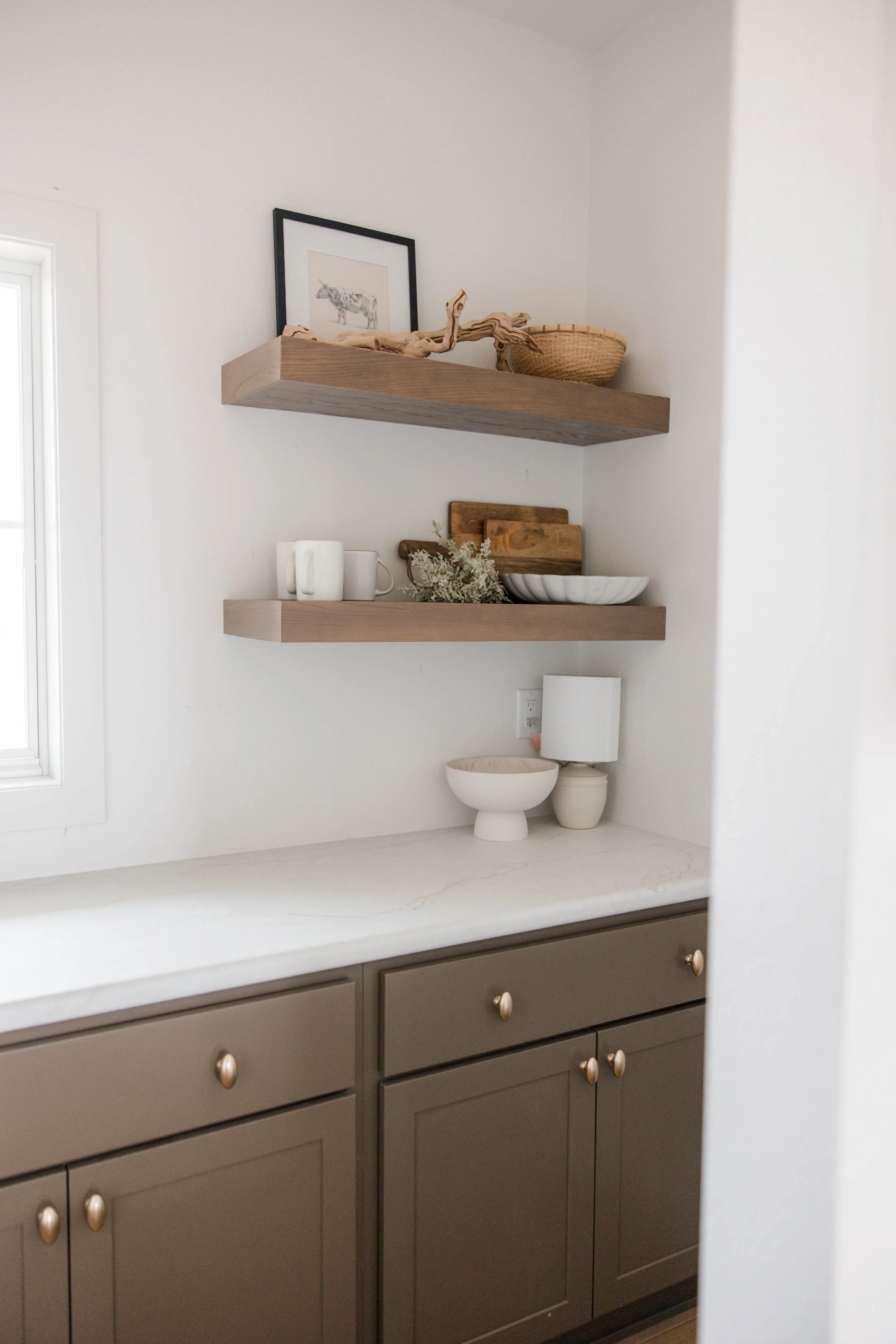 Kitchen window sill with gray cabinets, two white mugs, a white bowl, and decorative items including a small wooden cutting board, a white bowl, a basket, a framed picture of a zebra, a piece of driftwood, and dried greenery.