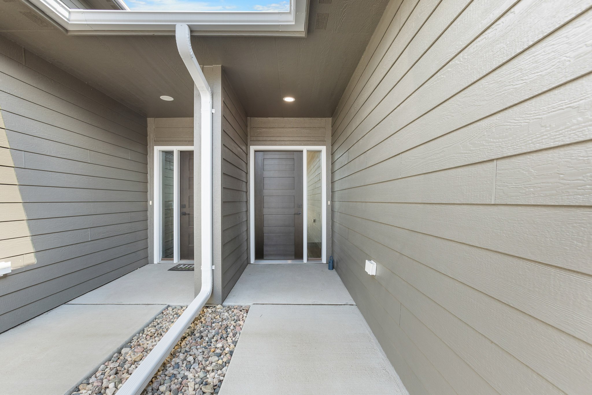 View of a small outdoor patio area with beige siding walls, a glass door, a white gutter downspout, and a small section of pebbles along the edge of the concrete floor.