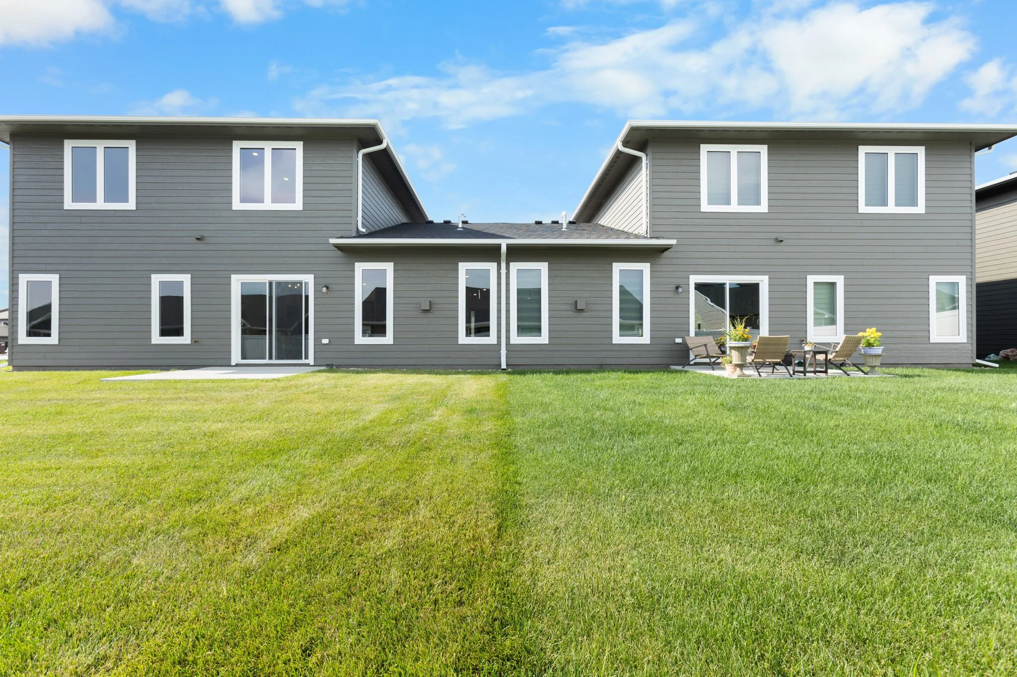 Two-story gray house with white trim, multiple windows, sliding glass door, and a small outdoor patio with chairs and potted plants in a grassy yard under a partly cloudy sky.
