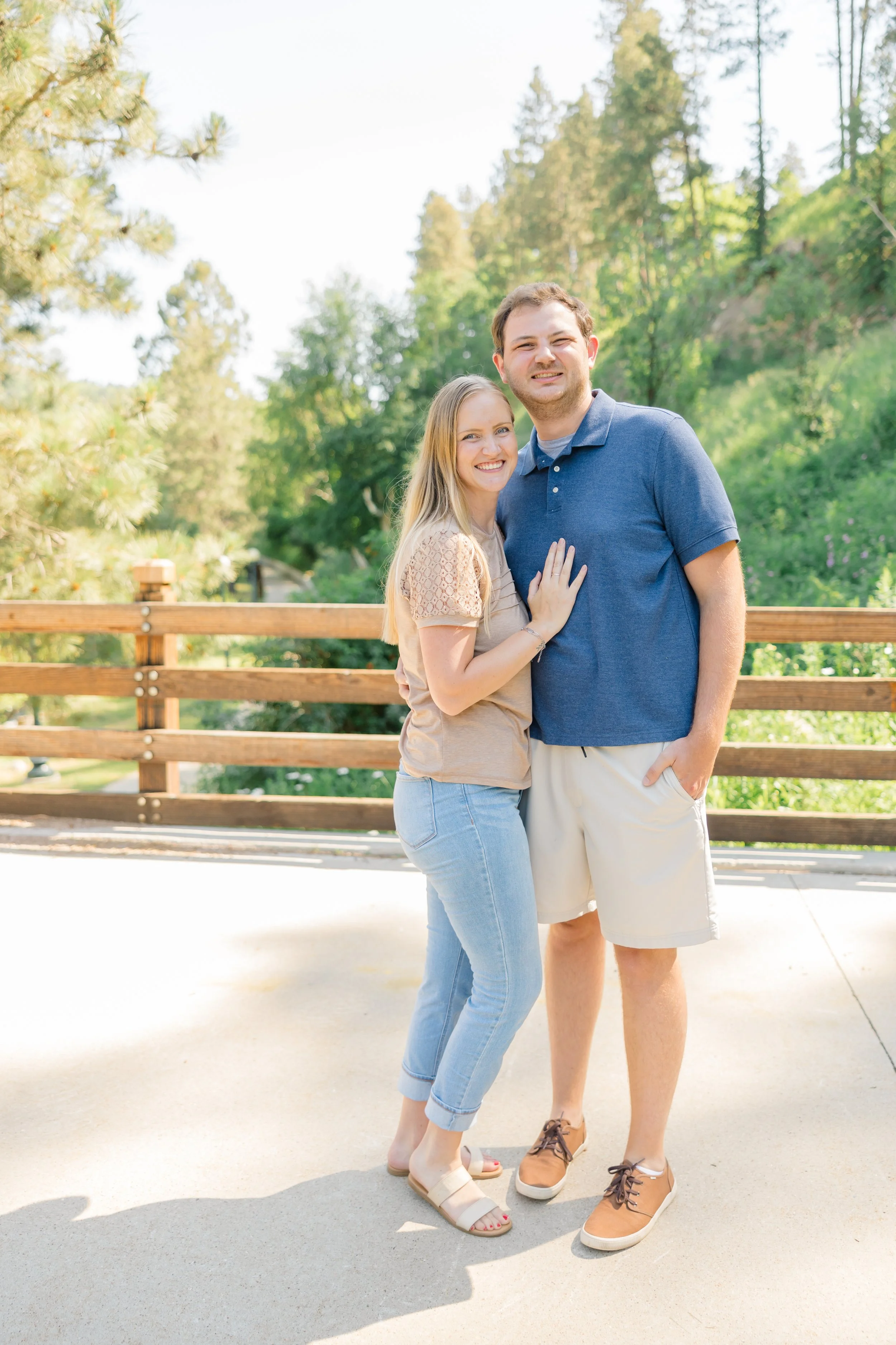 A smiling young woman with blonde hair and a smiling young man with brown hair standing close together outdoors on a bright sunny day, with a wooden fence and green trees in the background.