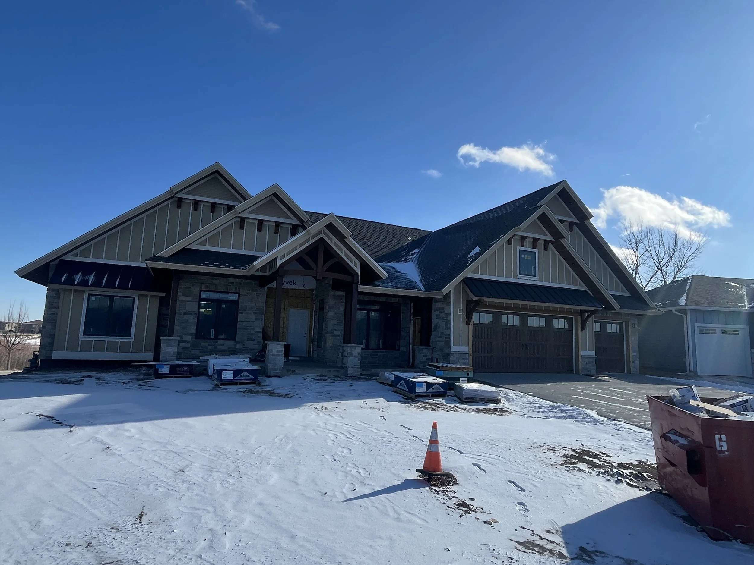 Under construction house with snow-covered front yard, construction materials, and construction cones, with a blue sky and few clouds.