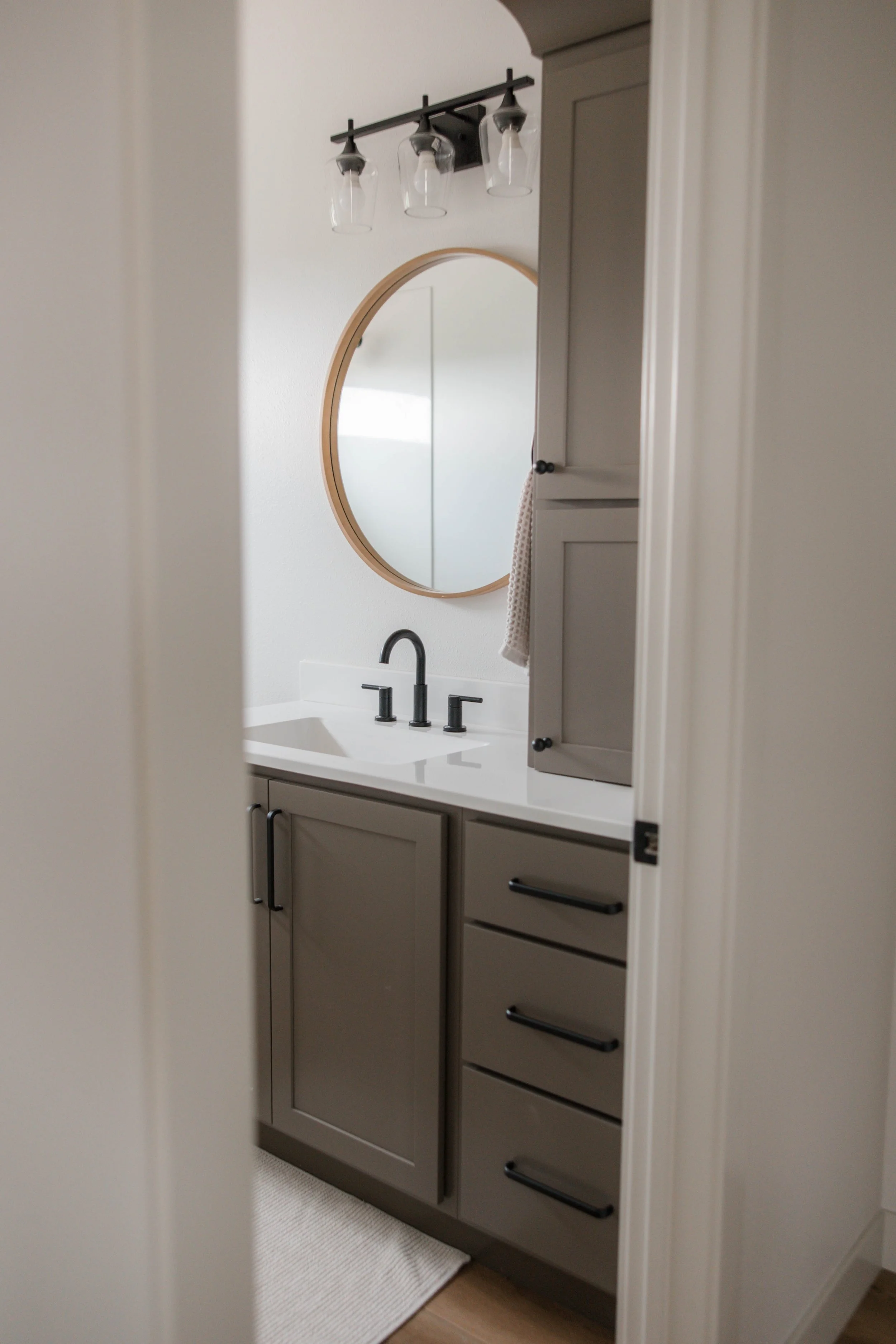 A bathroom vanity with a gray cabinet, a white countertop, a black faucet, an oval mirror, and a wall-mounted light fixture with three glass shades.