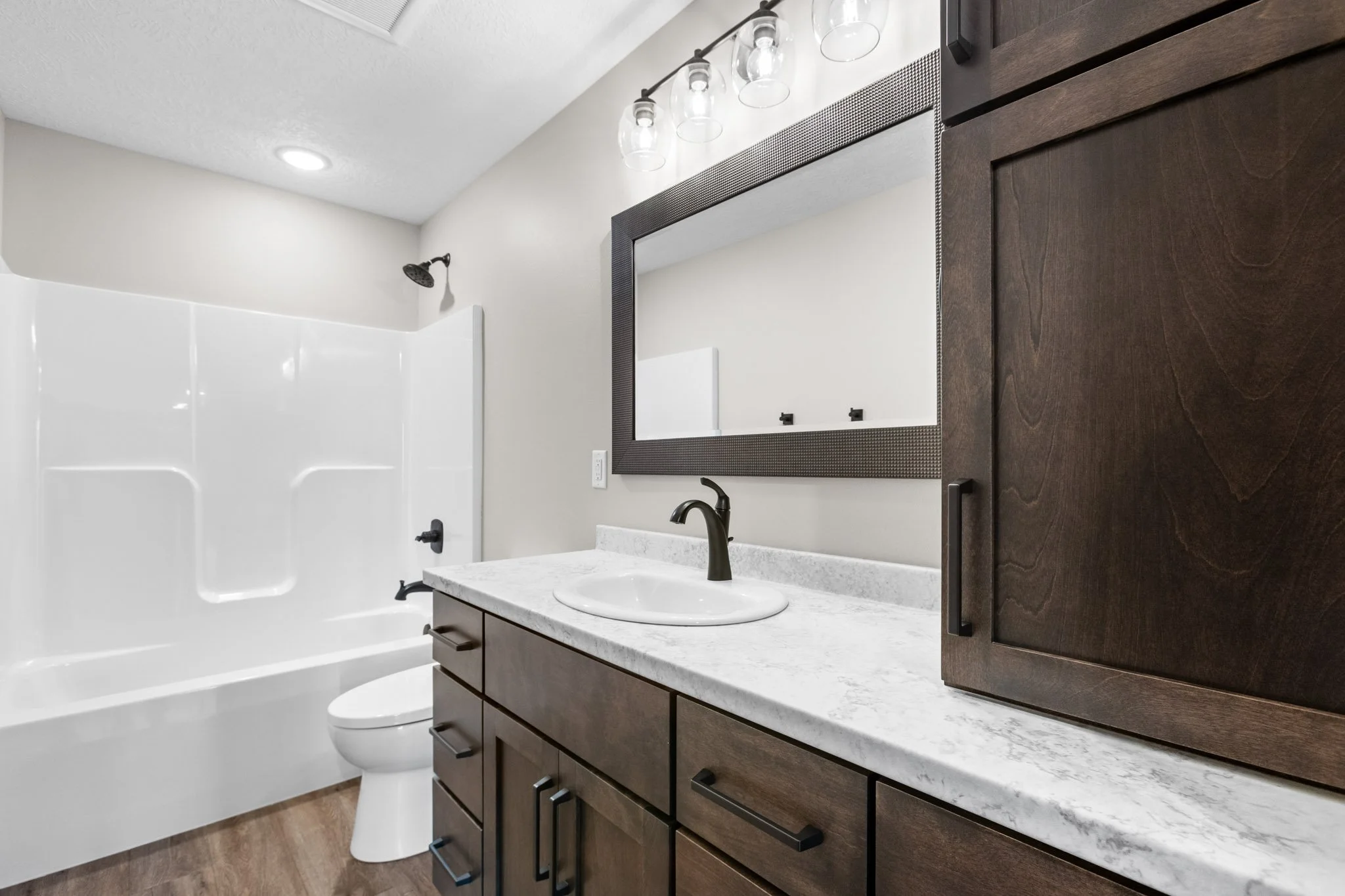 Modern bathroom with dark wood vanity, white countertop, oval sink, black faucet, large mirror, black light fixture, white bathtub with shower, beige walls, and wood flooring.