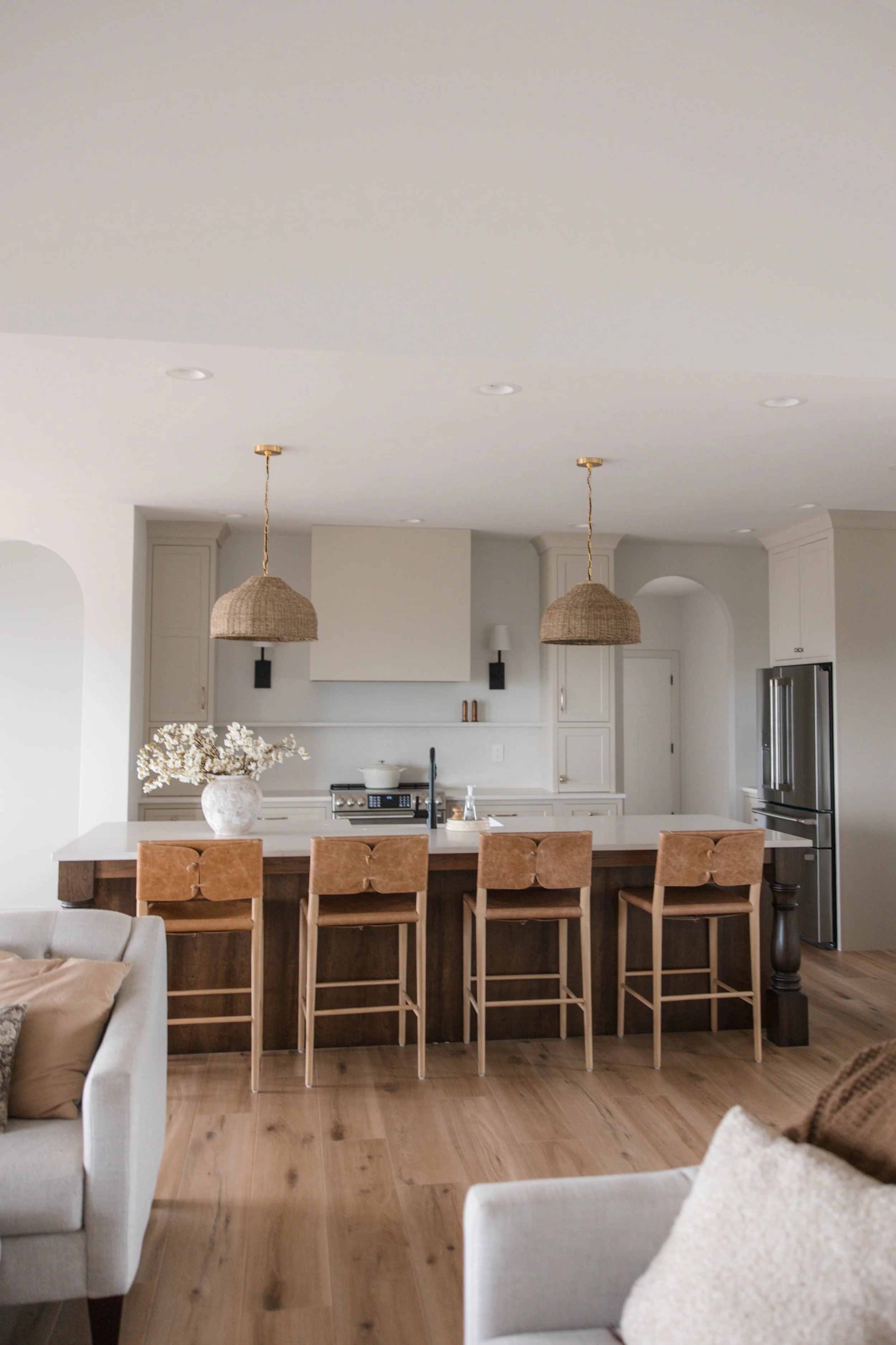 Modern open kitchen with light cabinets, a central island with a wooden countertop and four beige barstools, two hanging pendant lights with woven shades, a coffee machine, and a large stainless steel fridge in the background. There's a beige sofa wi