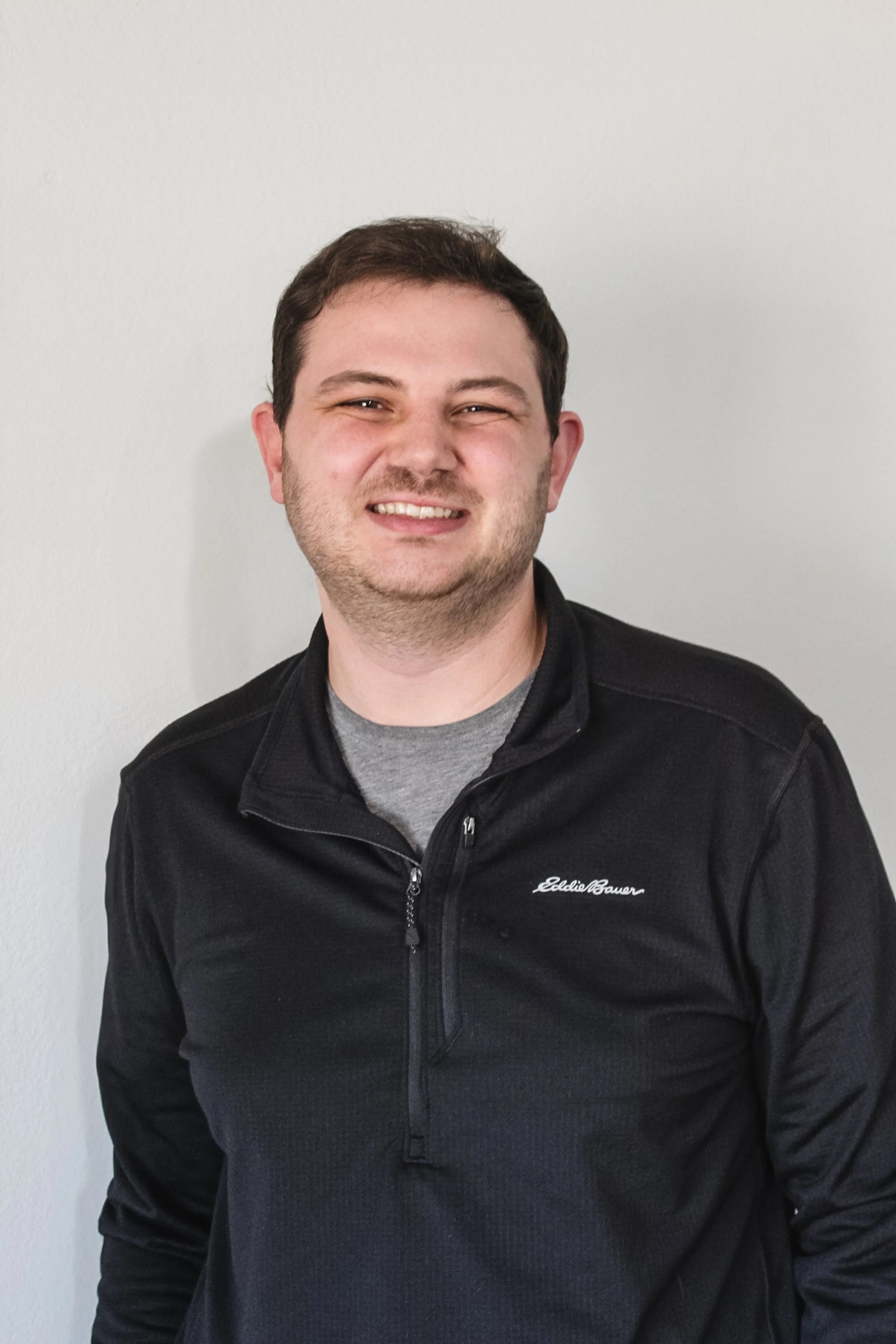 A young man smiling with short dark hair, wearing a black Eddie Bauer jacket and a gray t-shirt, standing against a plain white wall.