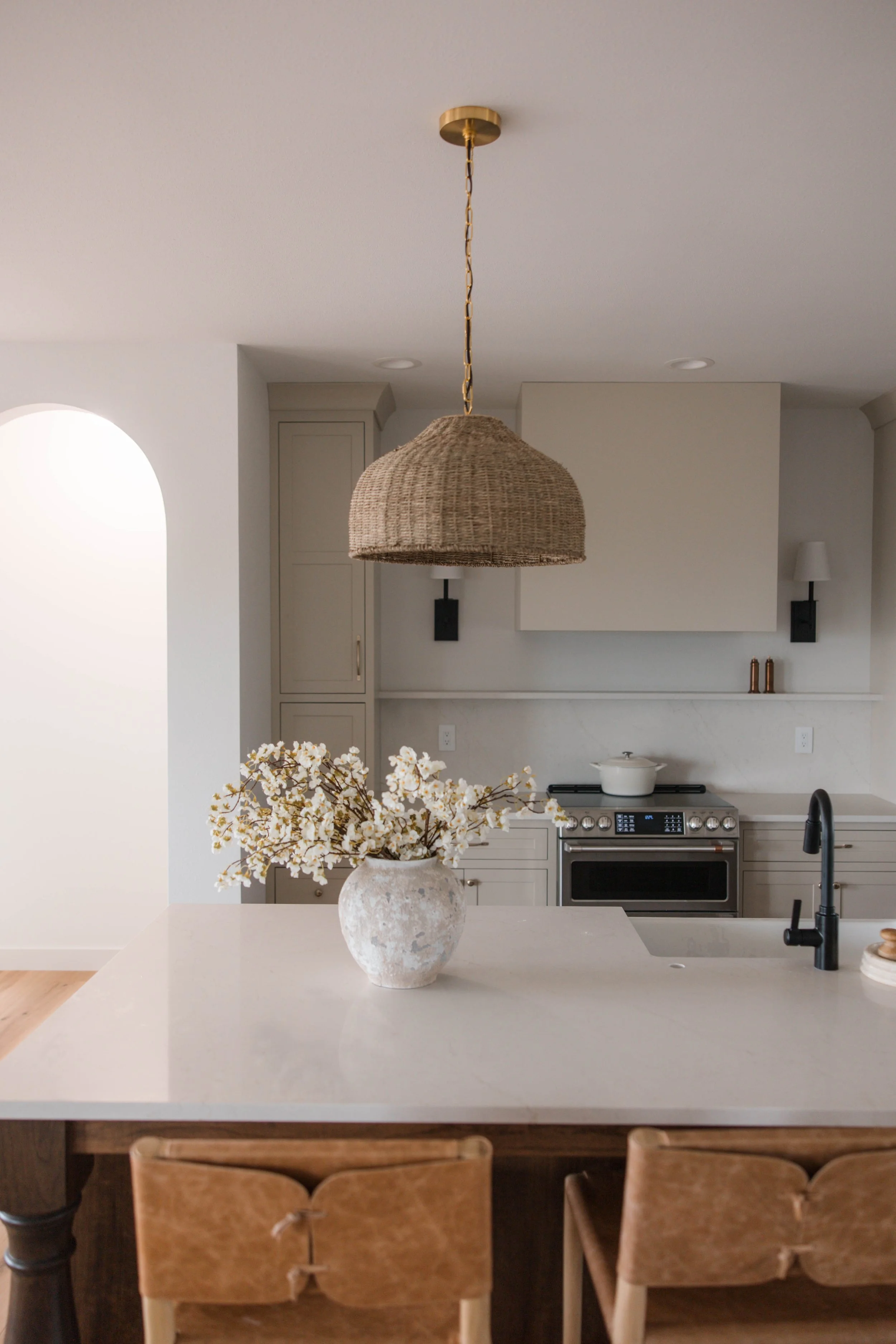 Modern kitchen with white countertop, beige cabinets, stainless steel stove, black backsplash, black faucet, and a vase of white flowers on the island. Pendant light hanging above.